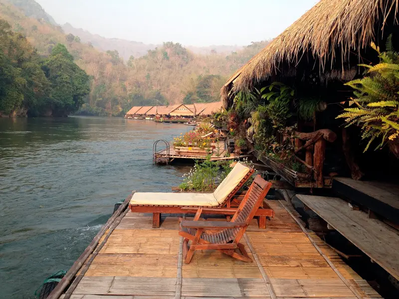 Zwei Holzliegen auf der Bambusterrasse eines Floßhotels am River Kwai inmitten der Dschungellandschaft Thailands.