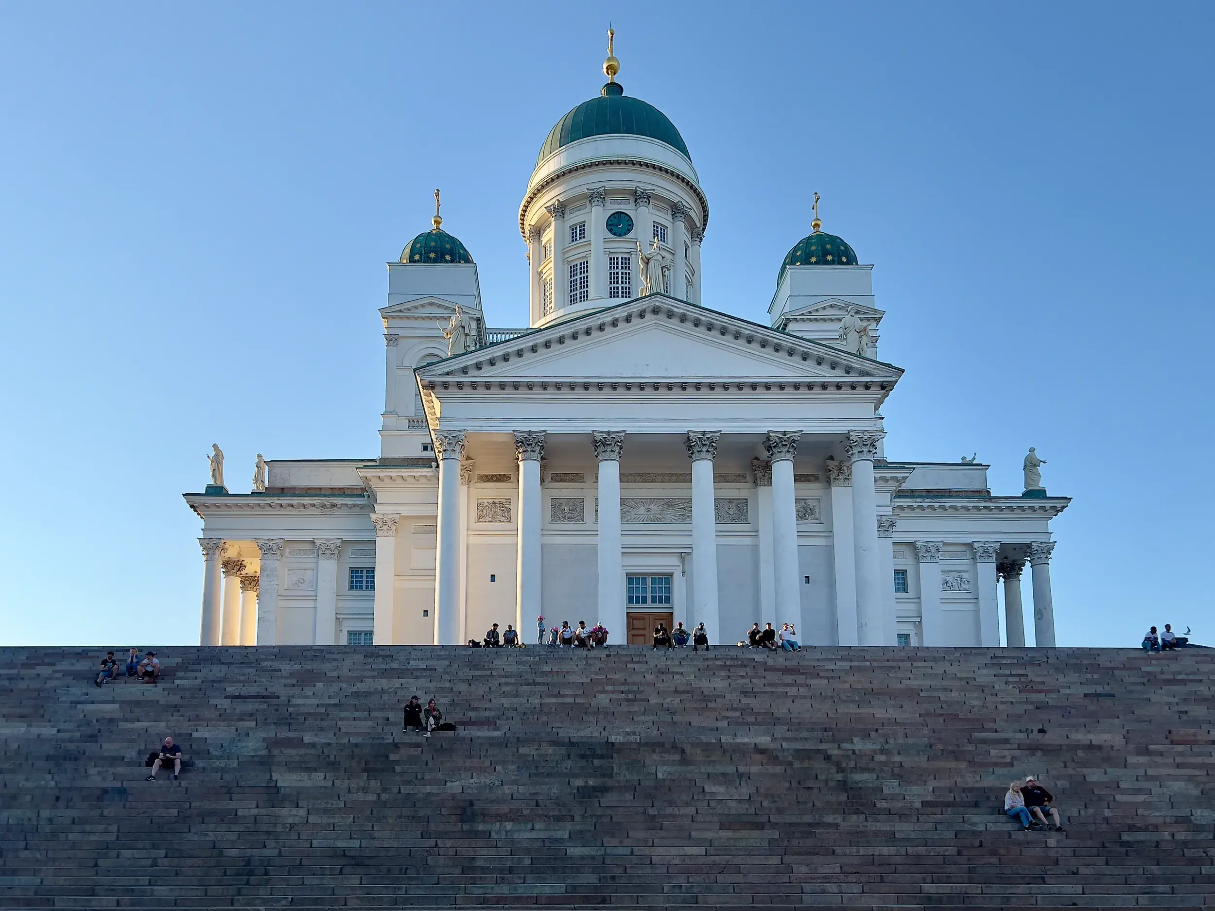 Strahlend weißer Dom von Helsinki mit grünen Kuppeln unter blauem Himmel, davor sitzen Menschen auf der breiten Steintreppe.