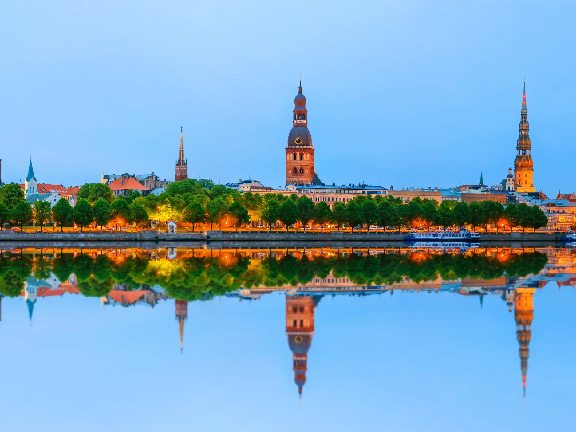 Die beleuchtete Skyline der Altstadt von Riga mit Dom und Petrikirche spiegelt sich in der Abenddämmerung im Fluss Düna.