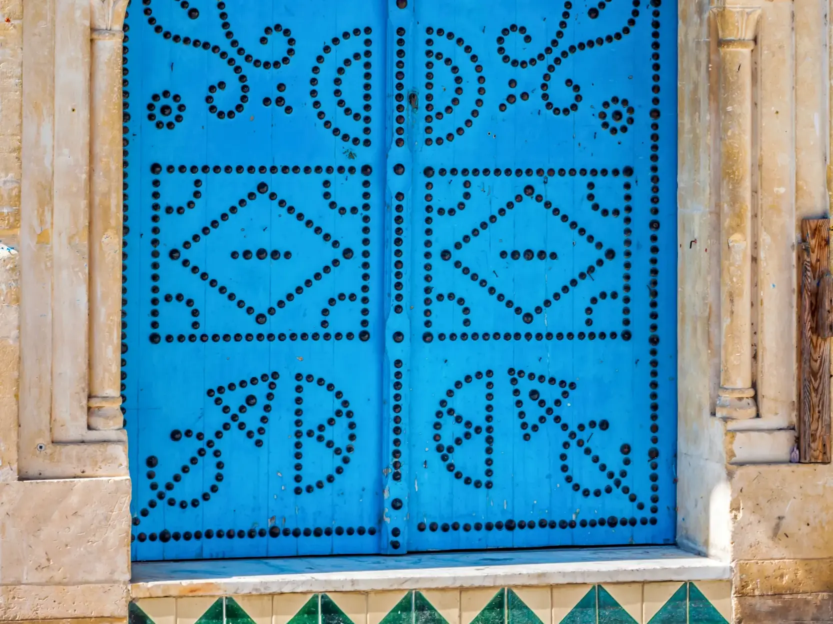 Kunstvoll mit schwarzen Nieten verzierte, azurblaue Holztür in einer hellen Steinmauer im Künstlerort Sidi Bou Said.