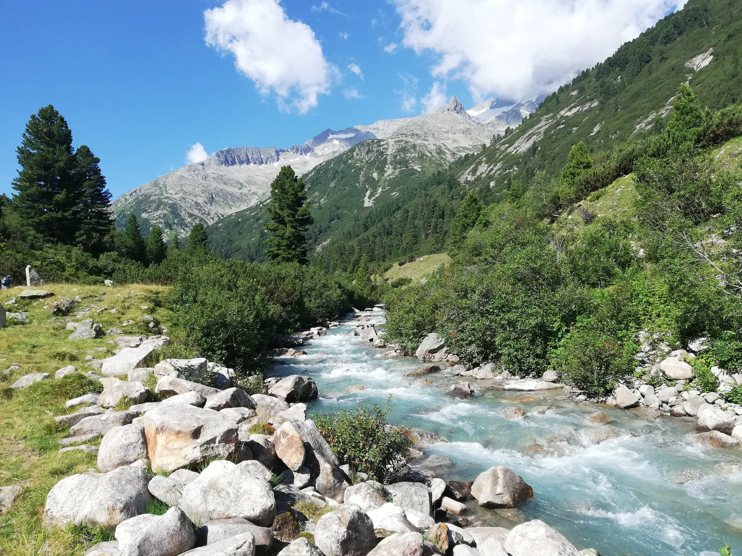 Türkisfarbener Bergbach rauscht auf der Alpenüberquerung durch das Pfitschtal vor der sonnigen Kulisse der Zillertaler Alpen.