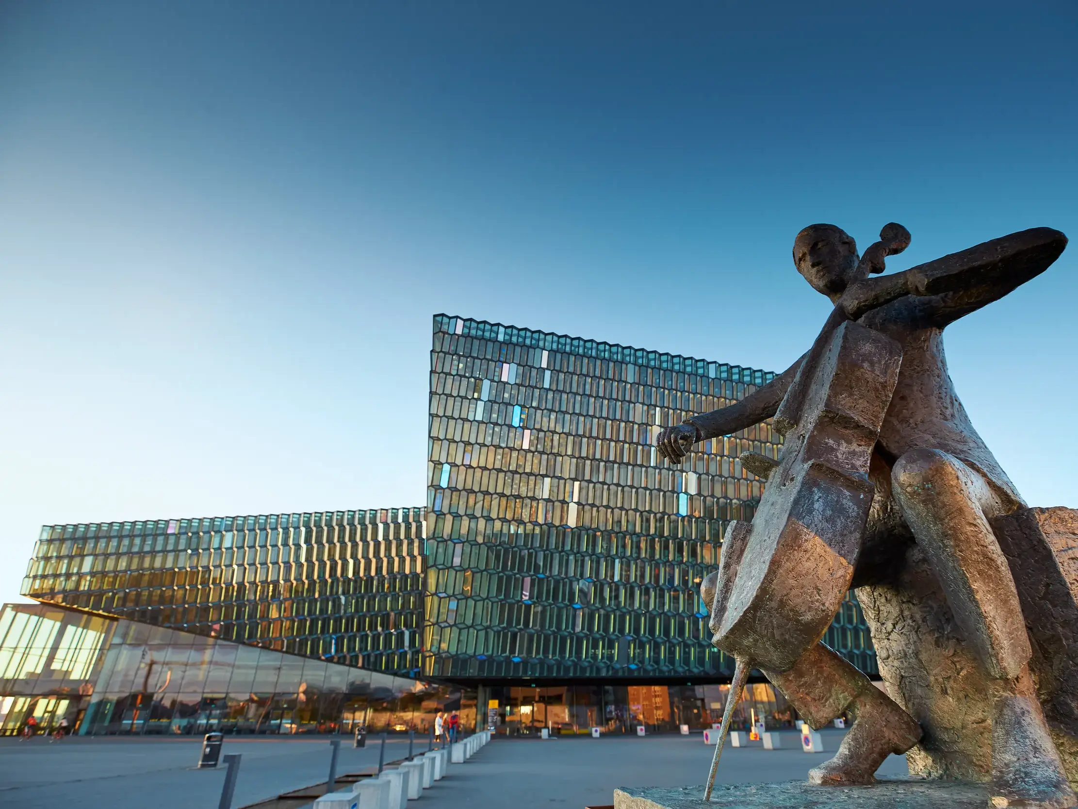 Bronzestatue eines Cellospielers vor der modernen Glasfassade eines Konzerthauses im Hafenviertel von Reykjavik, Island.