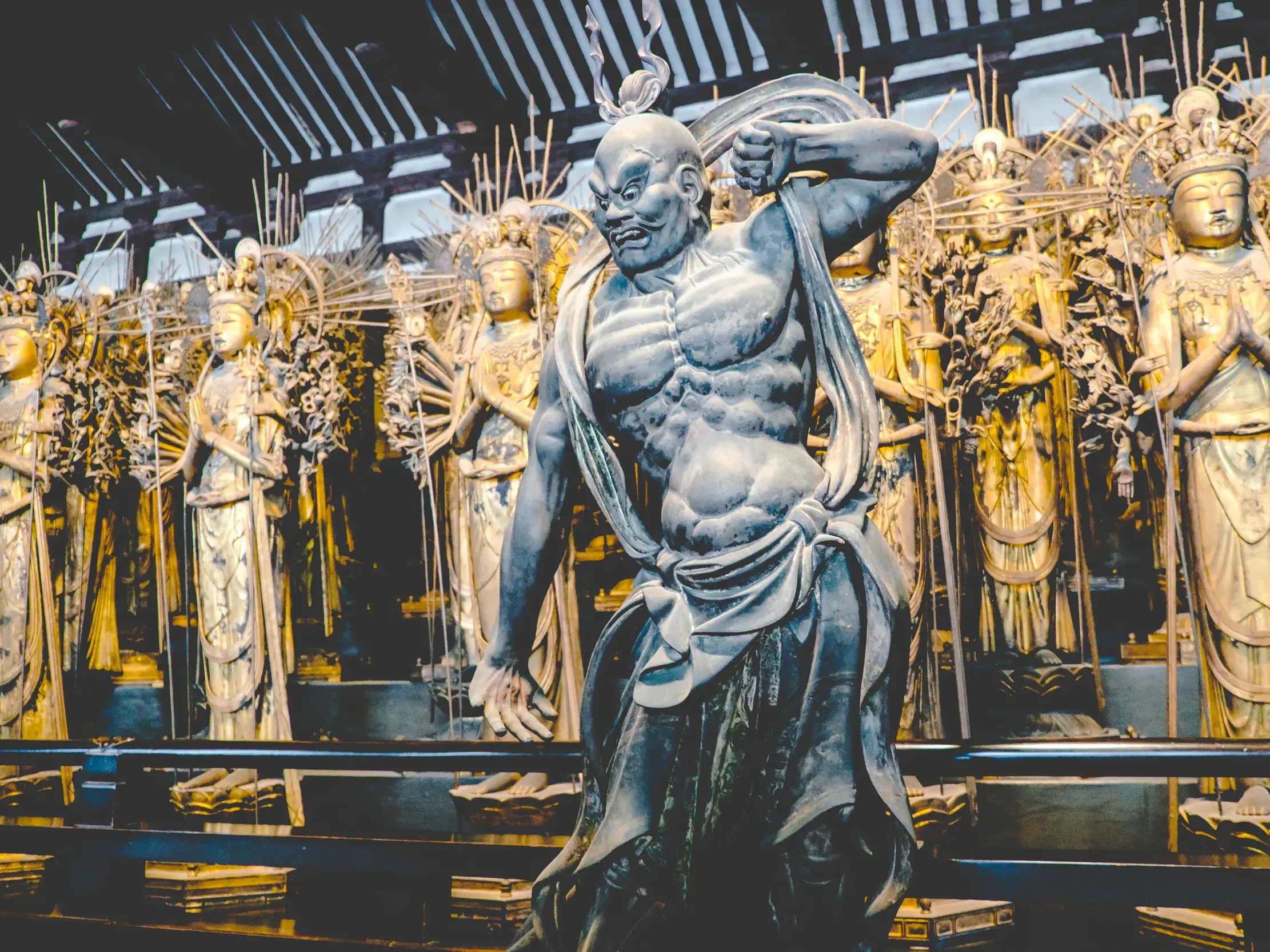 Muskulöse Wächterfigur in Kampfpose vor Reihen goldener Kannon-Statuen im Sanjusangendo-Tempel in Kyoto.