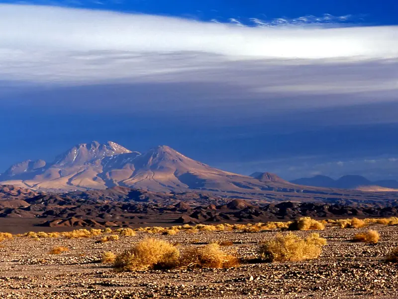 Weite, karge Wüstenlandschaft der Atacama in Chile mit Blick auf schneebedeckte Vulkane unter einem dramatischen Wolkenhimmel.