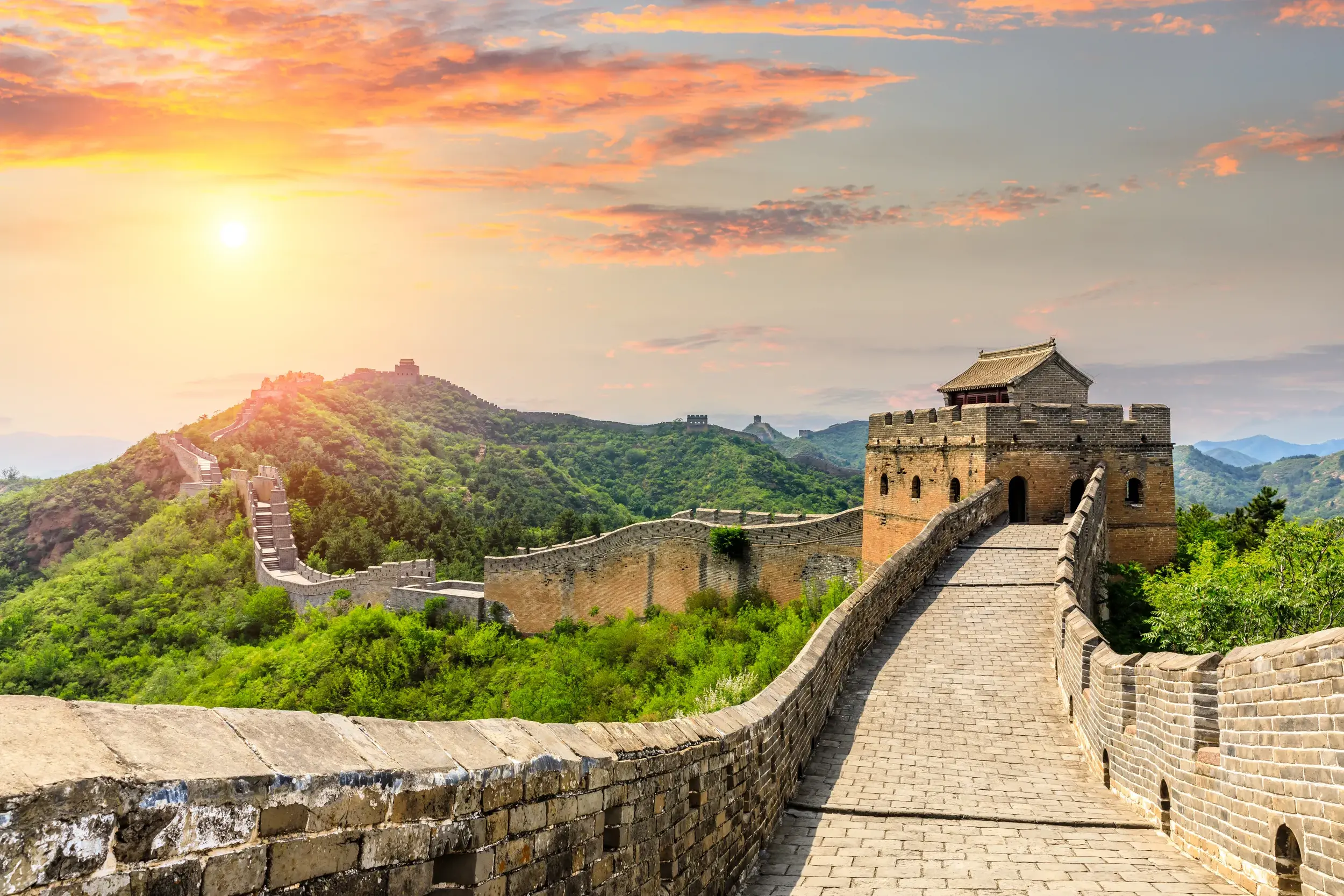 Ein Weg auf der Chinesischen Mauer bei Jinshanling führt bei Sonnenuntergang über grüne, bewaldete Hügel zu Wachtürmen.