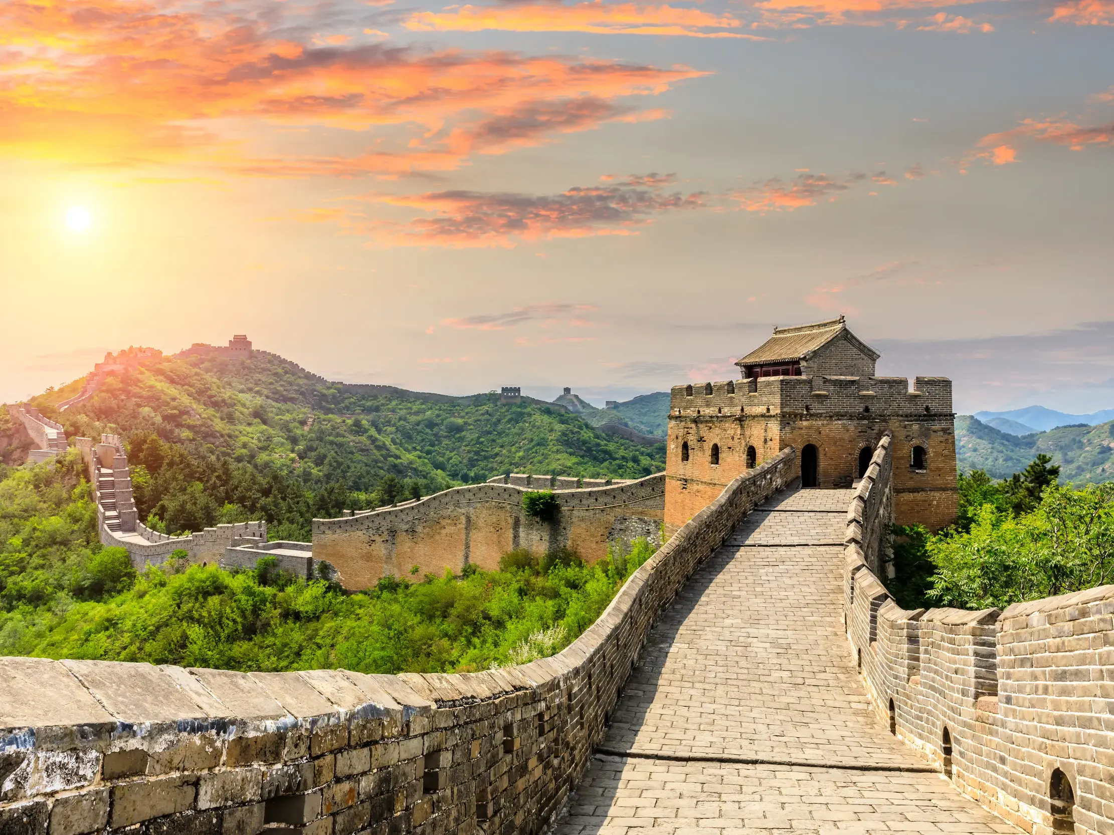Wanderweg auf der Chinesischen Mauer bei Jinshanling mit Wachturm und Blick über grüne Berge im Sonnenuntergang.