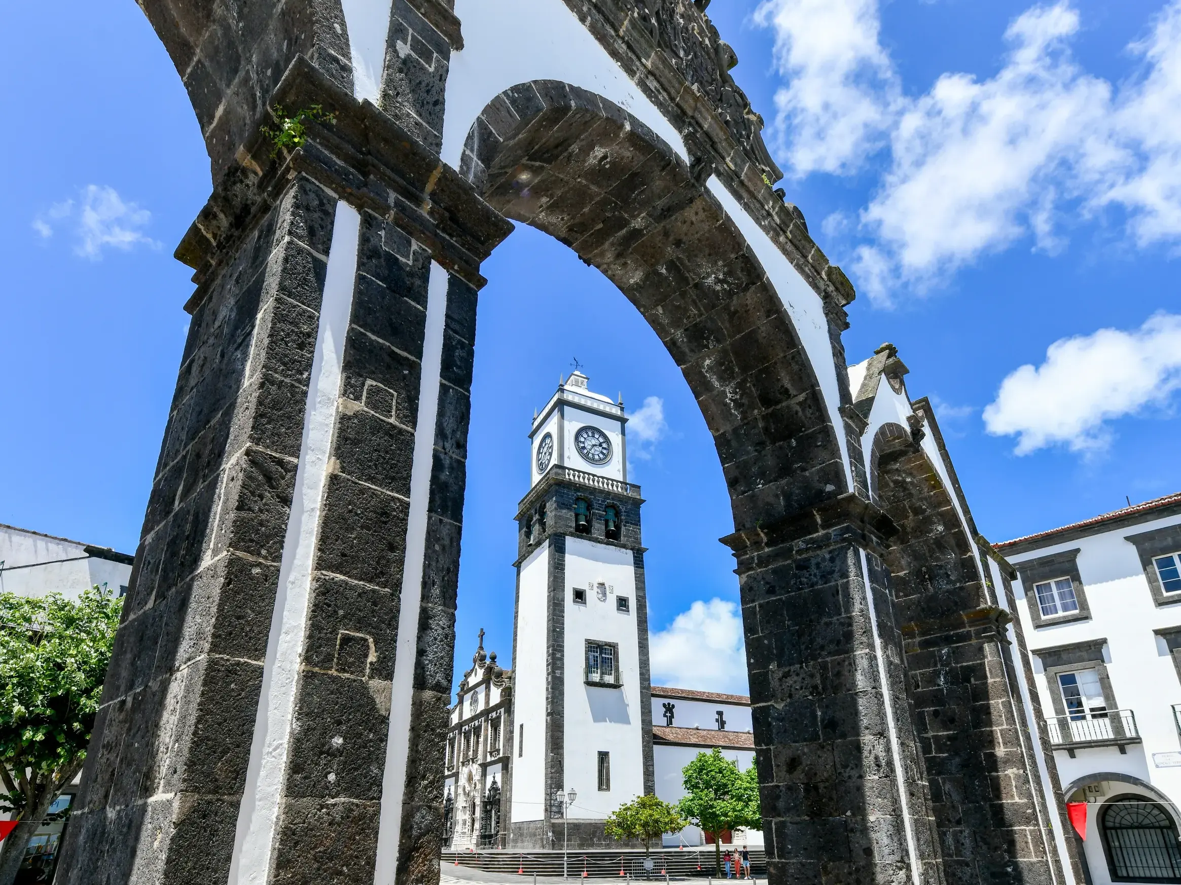 Das historische Stadttor aus Lavastein in Ponta Delgada, dahinter der Kirchturm vor blauem Himmel auf der Azoren-Insel São Miguel.