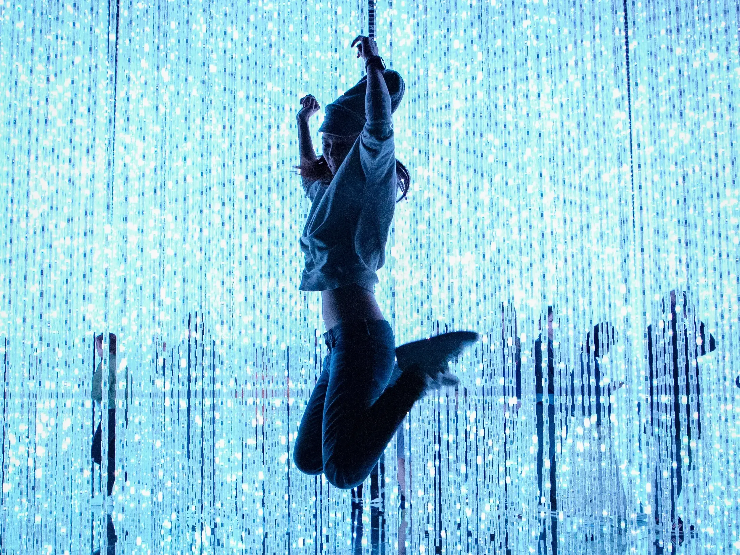 Begeisterte Besucherin springt in der immersiven Lichtinstallation des teamLab Museums in Tokyo zwischen blauen LED-Lichtsträngen.
