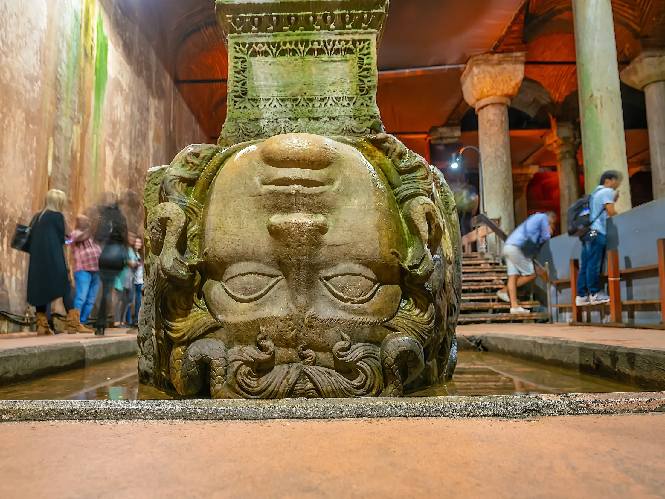 Umgedrehter Medusenkopf als Säulenbasis in der römischen unterirdischen Zisterne in Istanbul, mit Besuchern im Hintergrund.