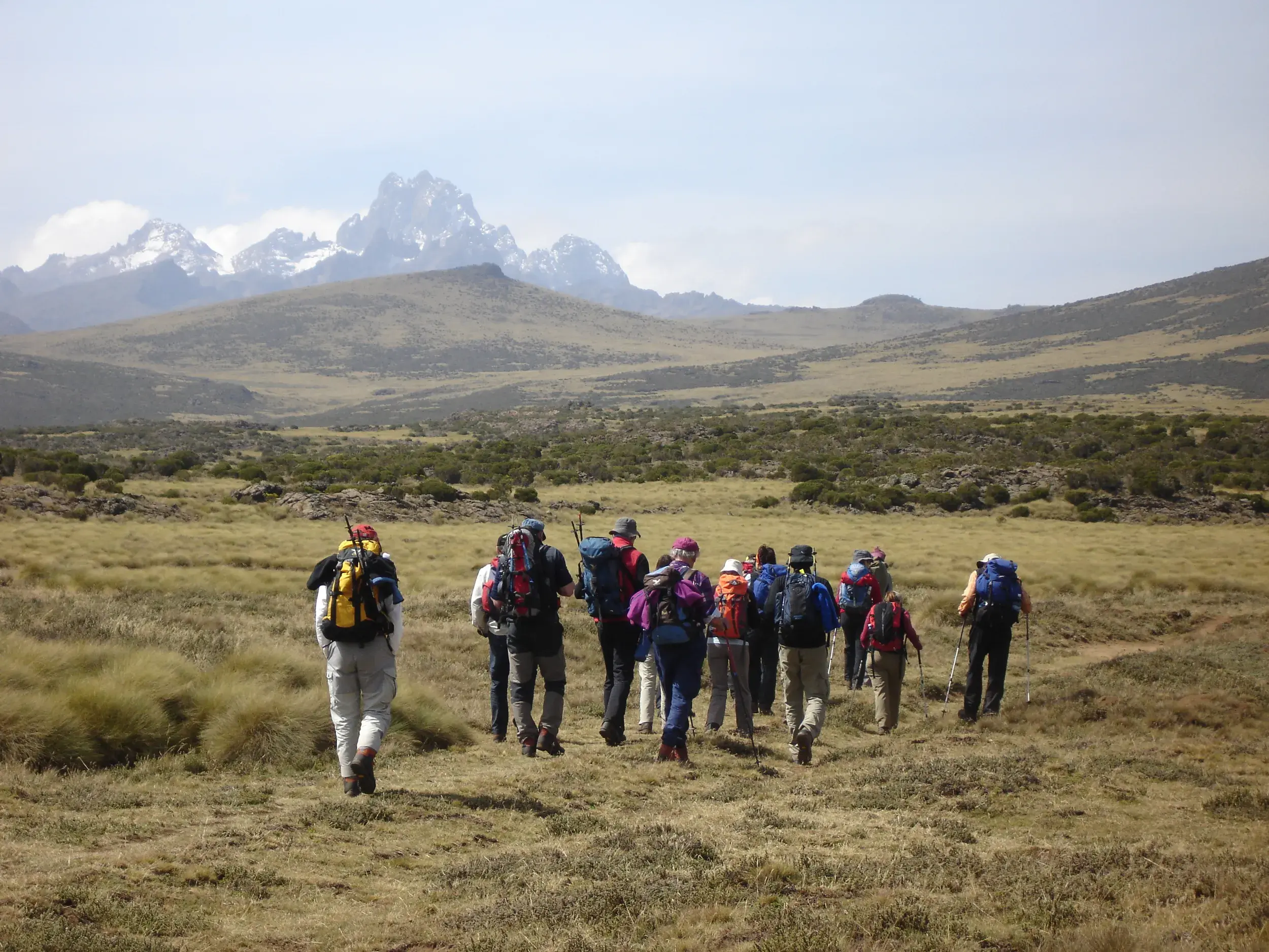 Eine Hauser-Wandergruppe mit Rucksäcken wandert durch eine weite Hochebene auf das schneebedeckte Mount-Kenya-Massiv zu.