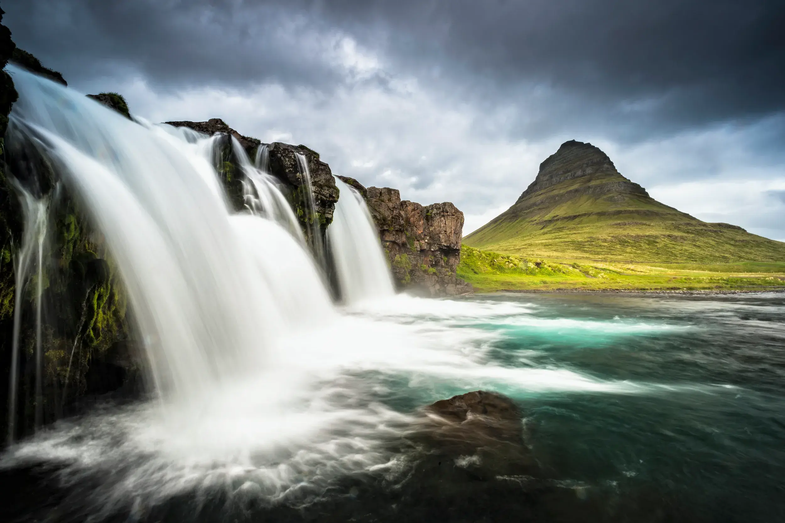 Mehrstufiger Wasserfall auf der Halbinsel Snaefellsnes in Island, im Hintergrund ein markanter grüner Berg unter dunklen Wolken.