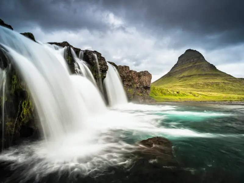 Mehrstufiger Wasserfall auf der Halbinsel Snaefellsnes in Island, im Hintergrund ein markanter grüner Berg unter dunklen Wolken.