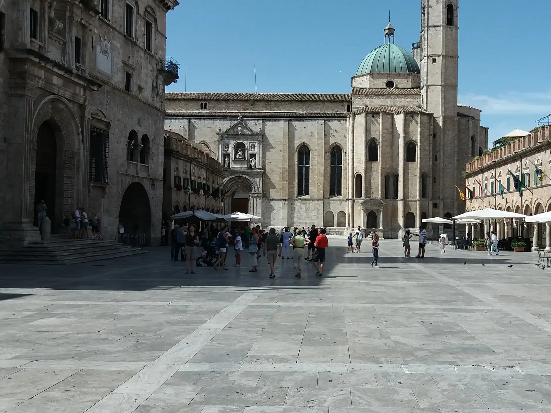 Besucher erkunden den sonnigen Piazza del Popolo in Ascoli Piceno, umgeben von historischen Gebäuden und Cafés.