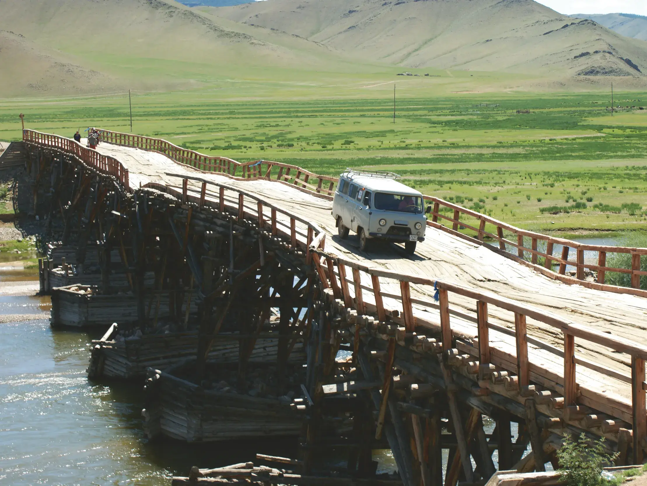 Begleitfahrzeug einer Radreise überquert eine rustikale Holzbrücke über einen Fluss in der mongolischen Steppe.