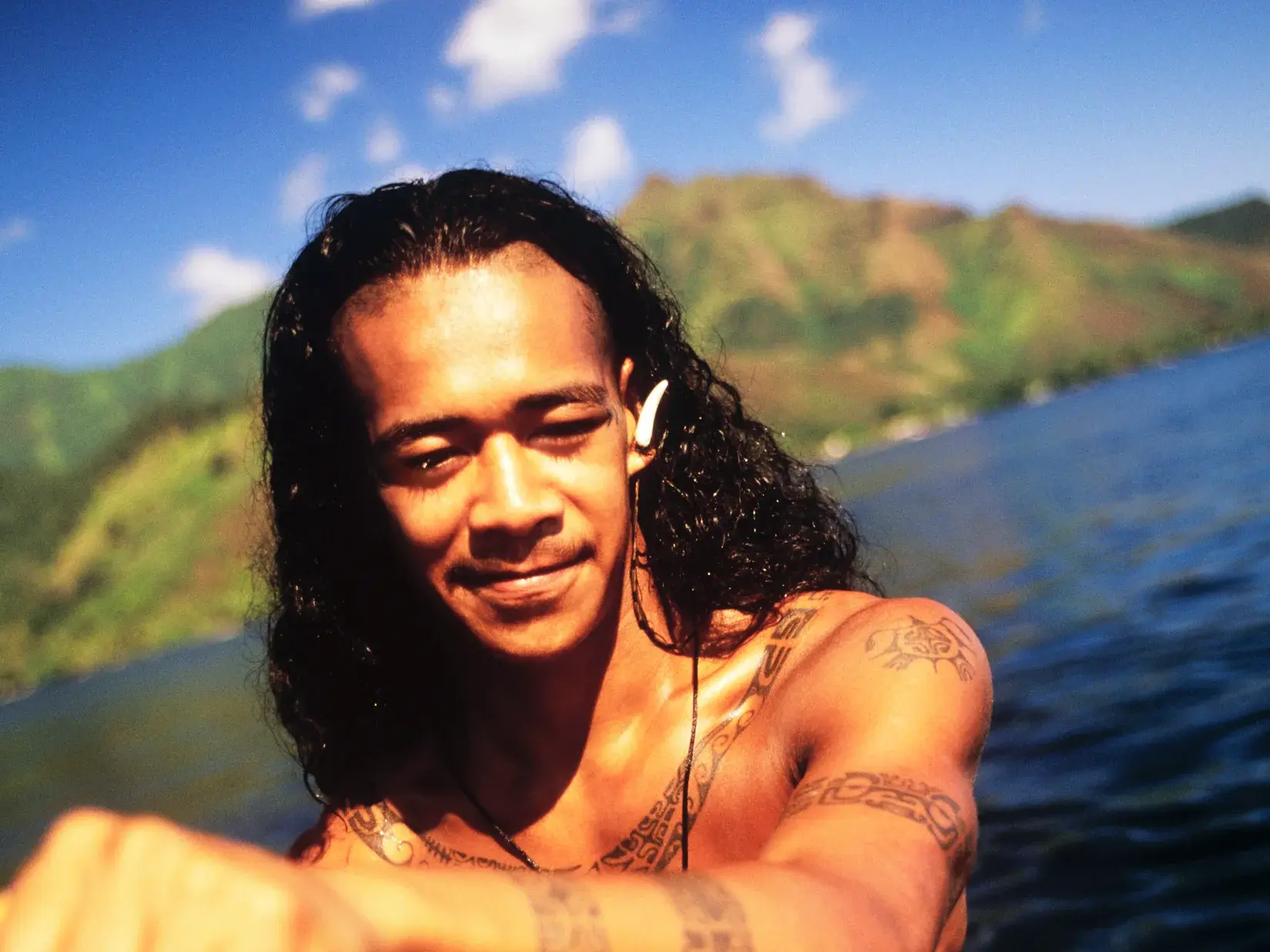 Einheimischer Mann mit traditionellen Tattoos und Muschelschmuck im Haar in einer Bucht vor den Vulkanbergen von Hawaii.