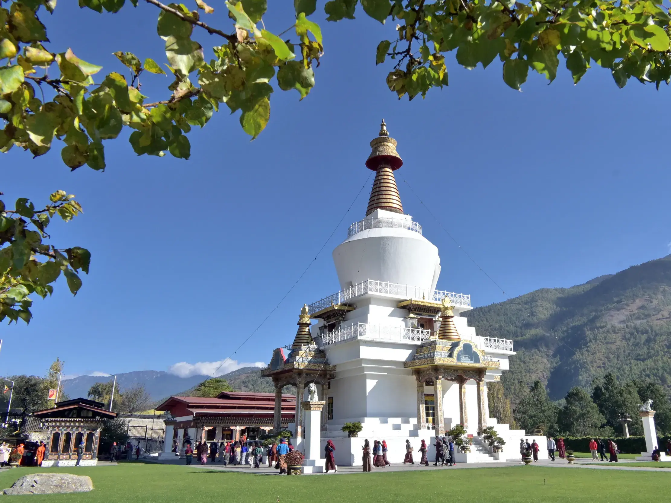 Gläubige und eine Reisegruppe umrunden den weißen König-Gedächtnis-Chörten in Thimphu vor einer Bergkulisse Bhutans.
