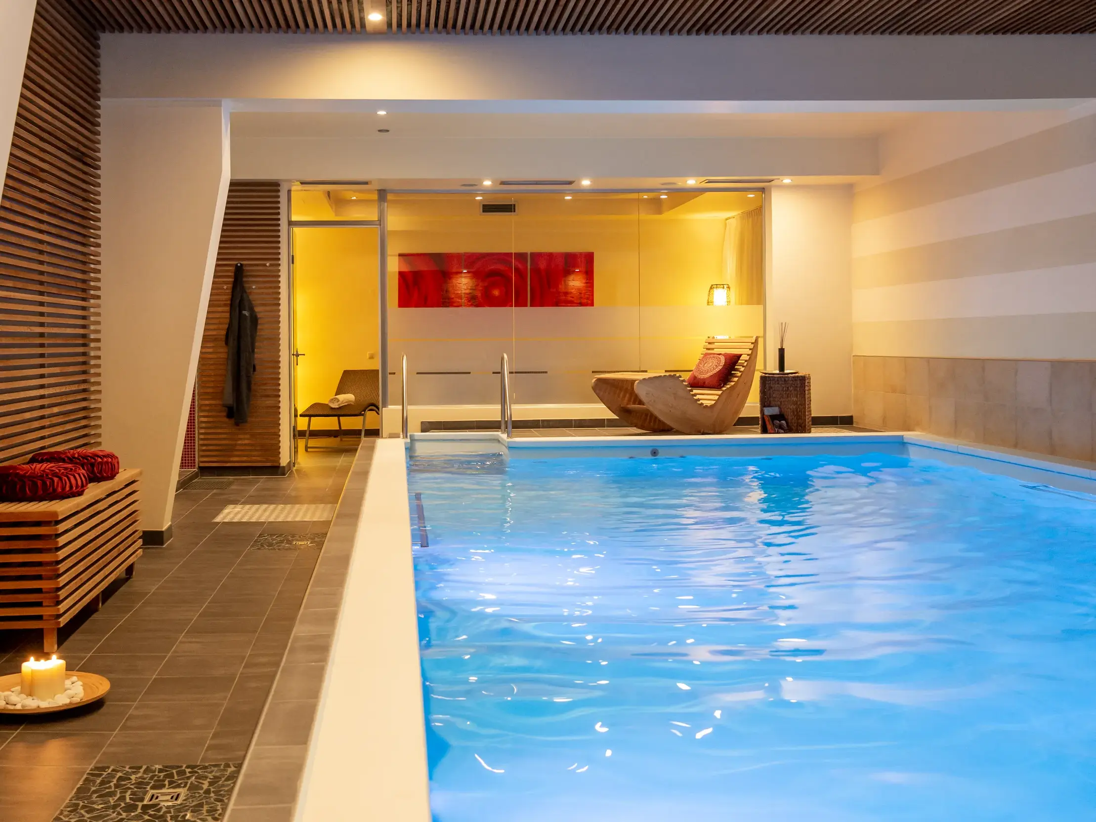 Moderner Hotel-Innenpool mit blau beleuchtetem Wasser und angrenzendem Ruhebereich mit Holzliege und warmem Licht.