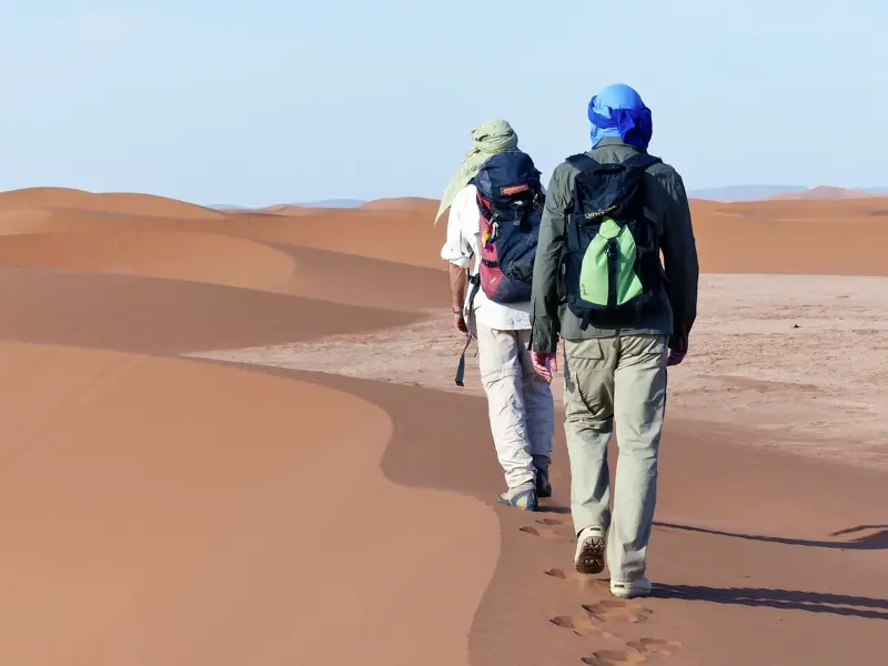 Zwei Trekking-Reisende wandern mit Rucksäcken durch die weiten, rötlichen Sanddünen der Sahara in Marokko.