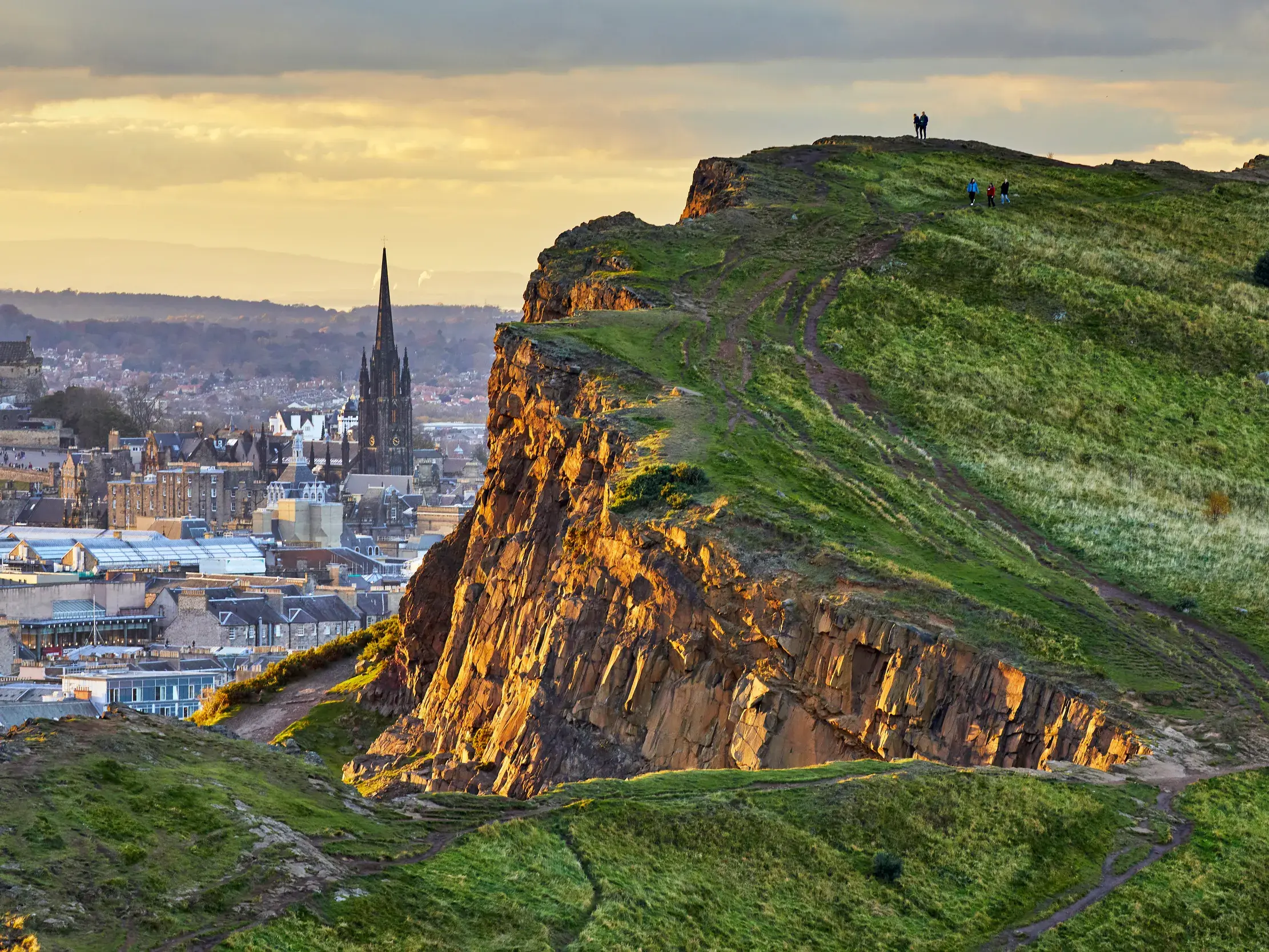 Wanderer auf einem grünen, felsigen Hügel genießen den Ausblick auf die Skyline von Edinburgh in der goldenen Abendsonne.