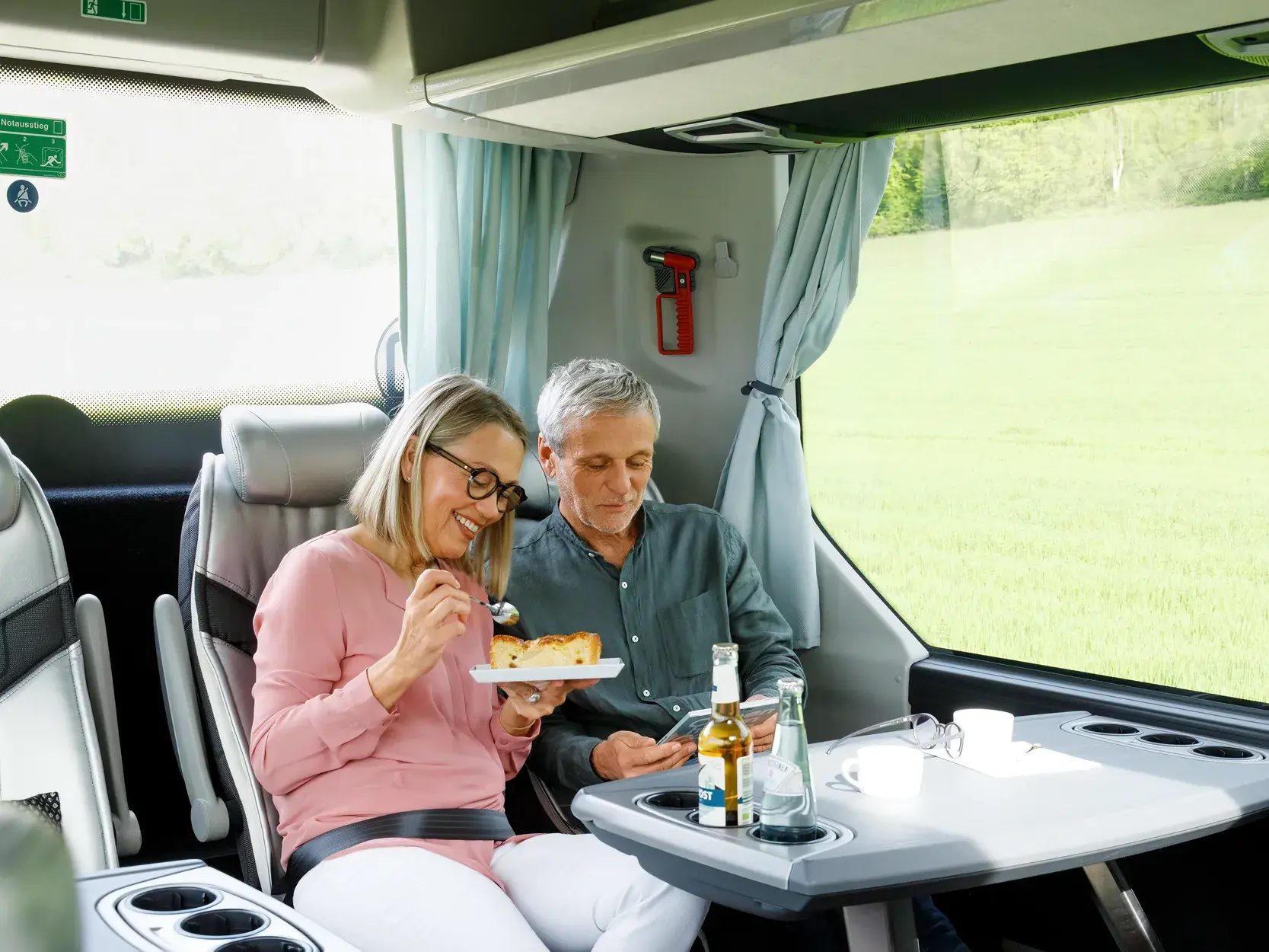 Lächelndes Paar auf einer Studiosus-Studienreise genießt einen Imbiss im Komfortbus mit Blick auf eine vorbeiziehende Landschaft.