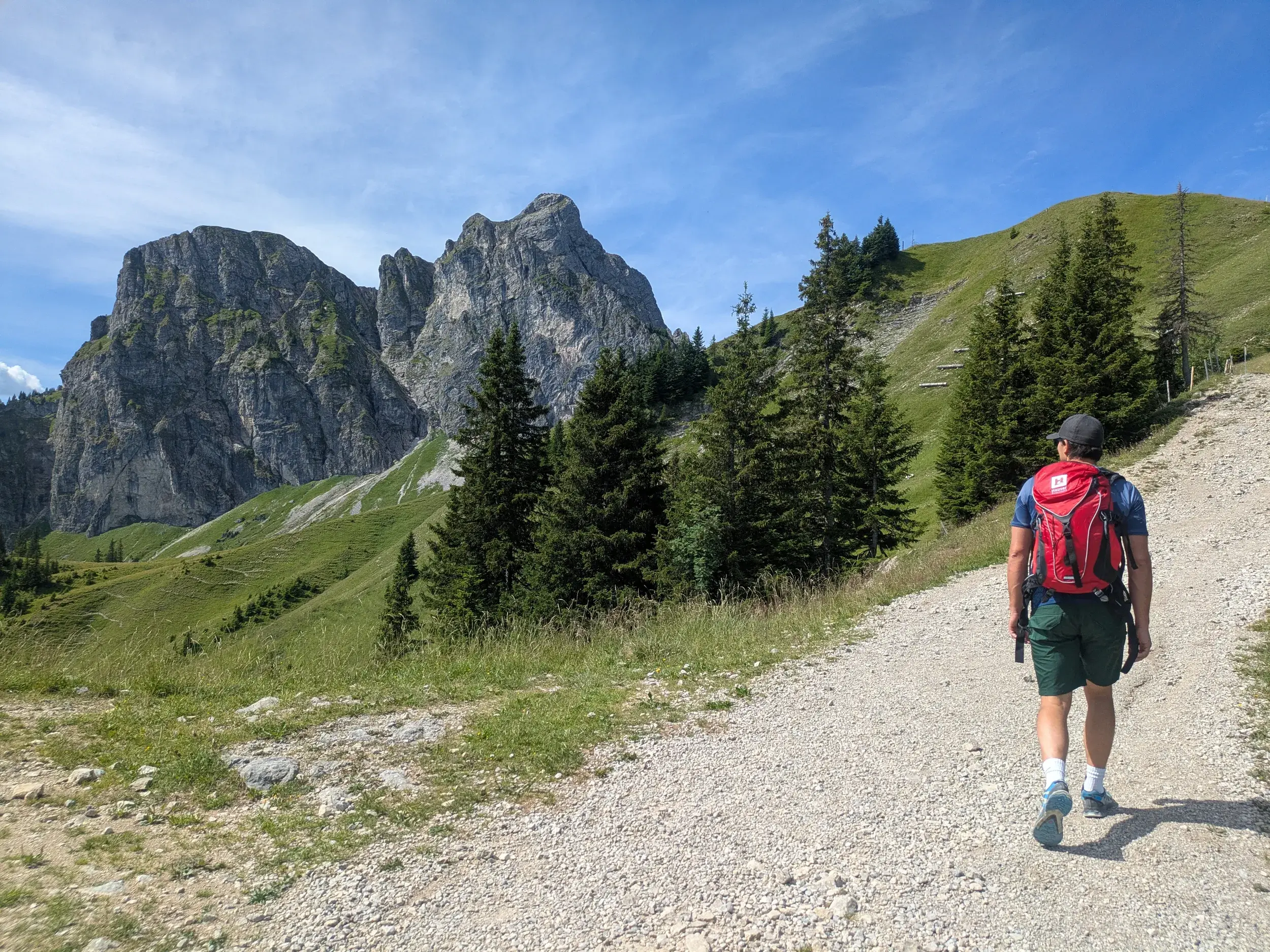 Ein Wanderer mit rotem Rucksack auf einem Schotterweg beim Aufstieg zum markanten Gipfel des Aggenstein im Ostallgäu.