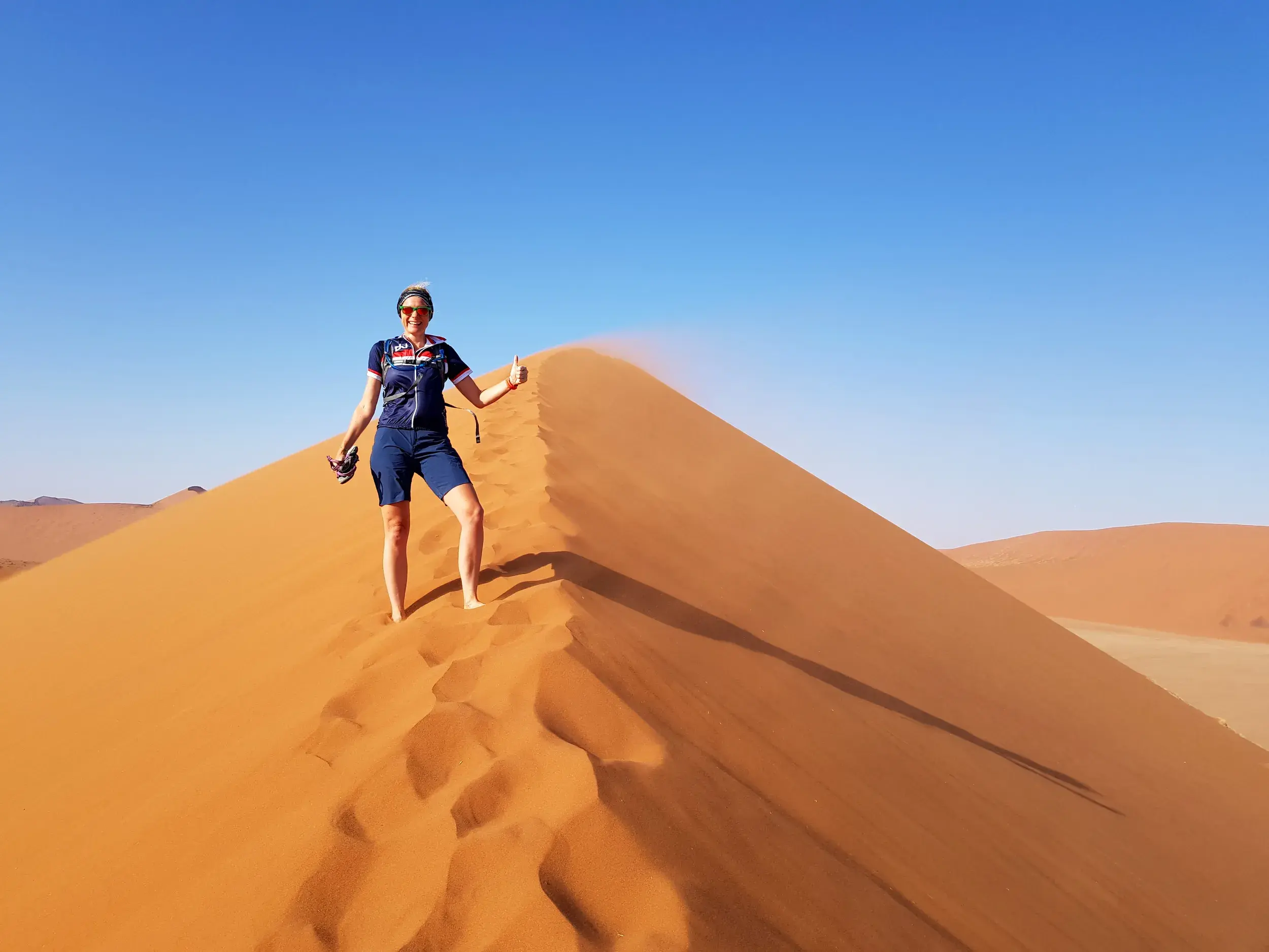 Eine Radfahrerin steht nach dem Aufstieg lächelnd auf einem Dünenkamm in der Namib-Wüste und gibt ein Daumen-hoch-Zeichen.