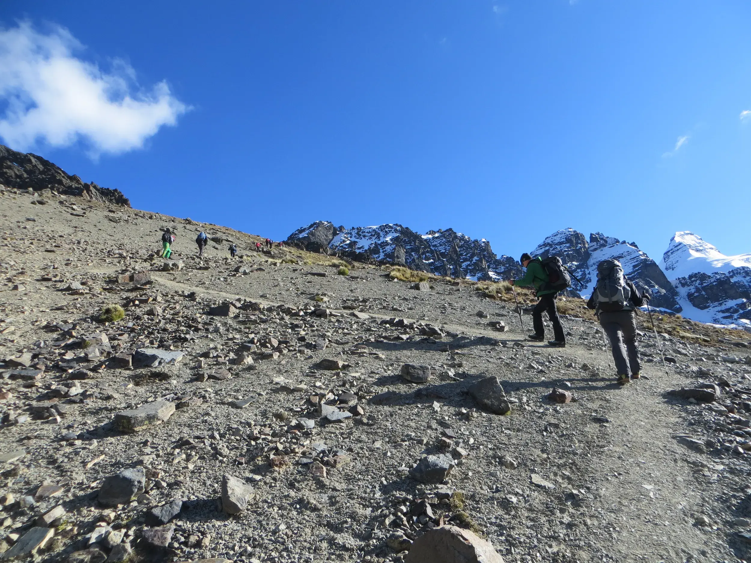 Eine Trekking-Gruppe steigt auf einem Geröllpfad im Condoriri Gebirge auf, im Hintergrund ragen schneebedeckte Andengipfel.