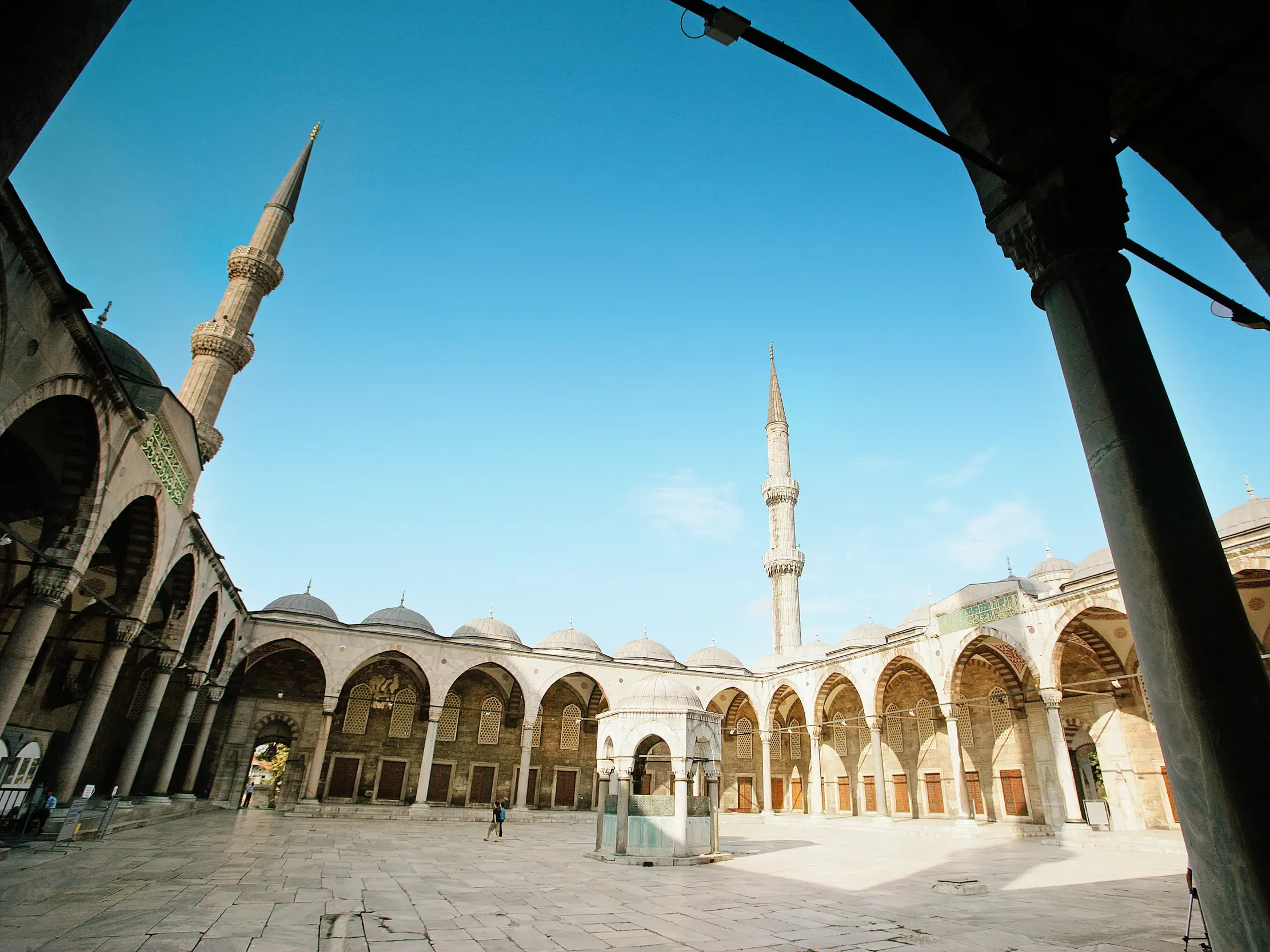 Weitläufiger Innenhof der Blauen Moschee in Istanbul mit steinernen Arkaden, Kuppeln und hohen Minaretten unter strahlend blauem Himmel.