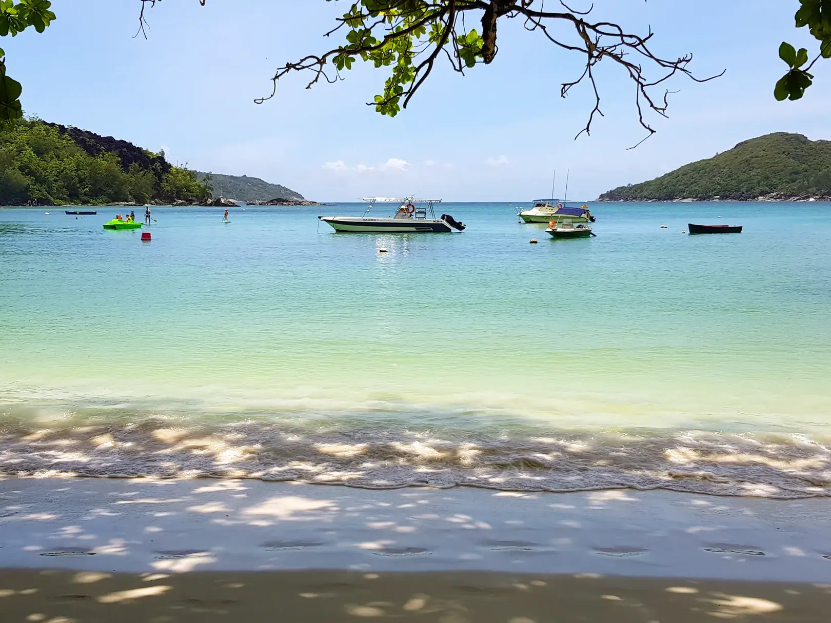 Kleine Boote ankern in einer idyllischen Bucht der Seychellen mit türkisfarbenem Wasser und bewaldeten Hügeln im Hintergrund.