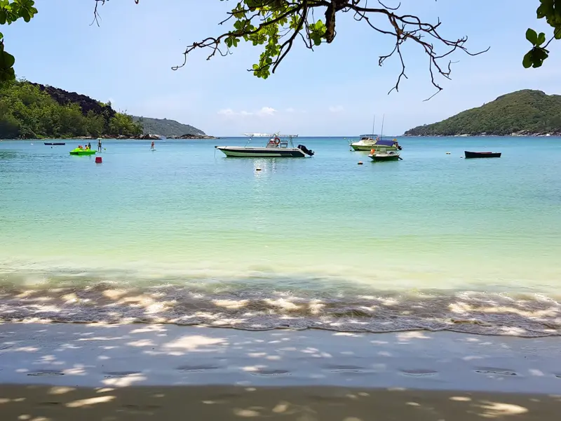 Kleine Boote ankern in einer idyllischen Bucht der Seychellen mit türkisfarbenem Wasser und bewaldeten Hügeln im Hintergrund.