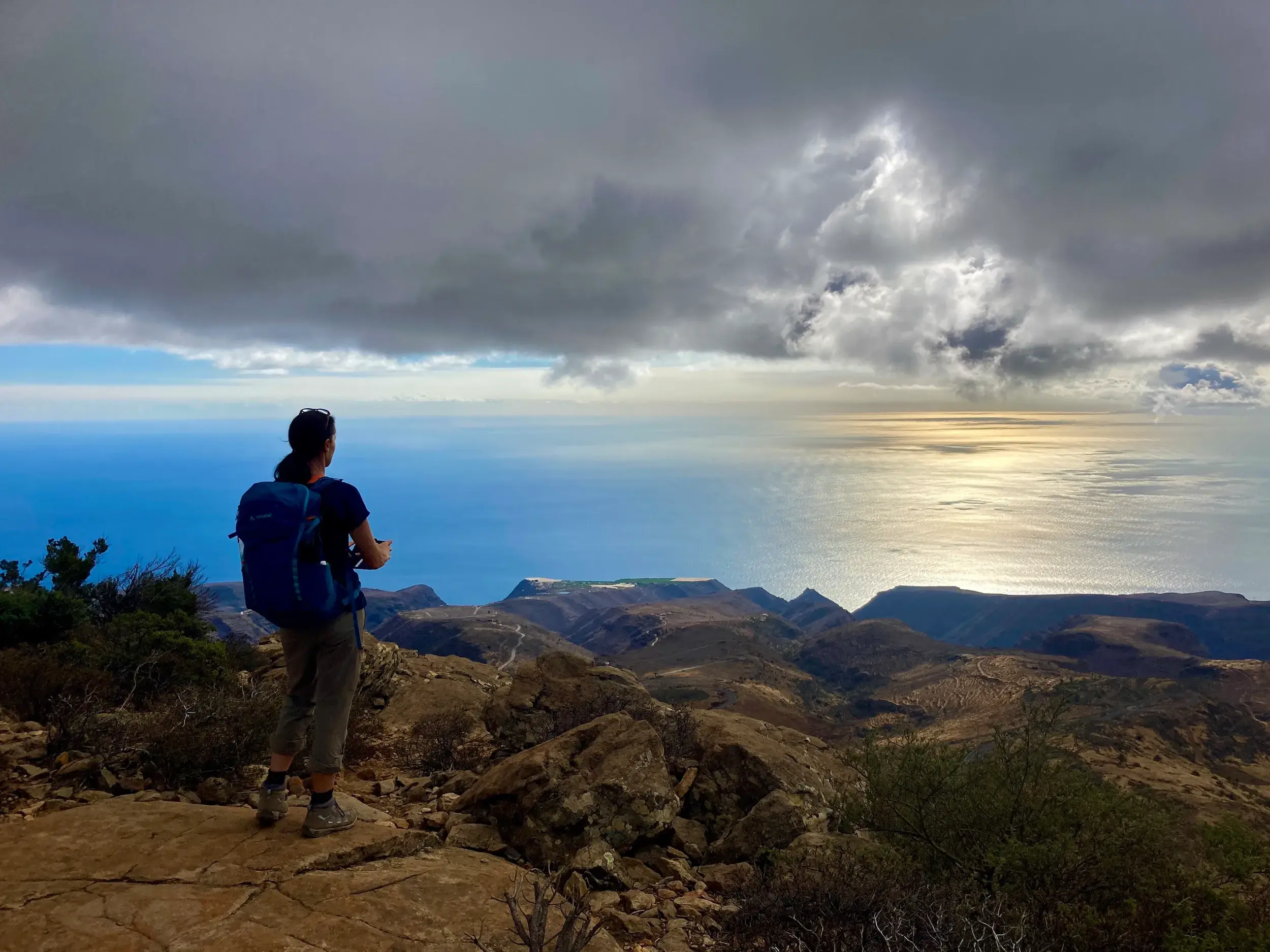 Eine Wanderin mit Rucksack genießt auf La Gomera die Aussicht von einem Felsen über die Berglandschaft auf den sonnenbeschienenen Atlantik.