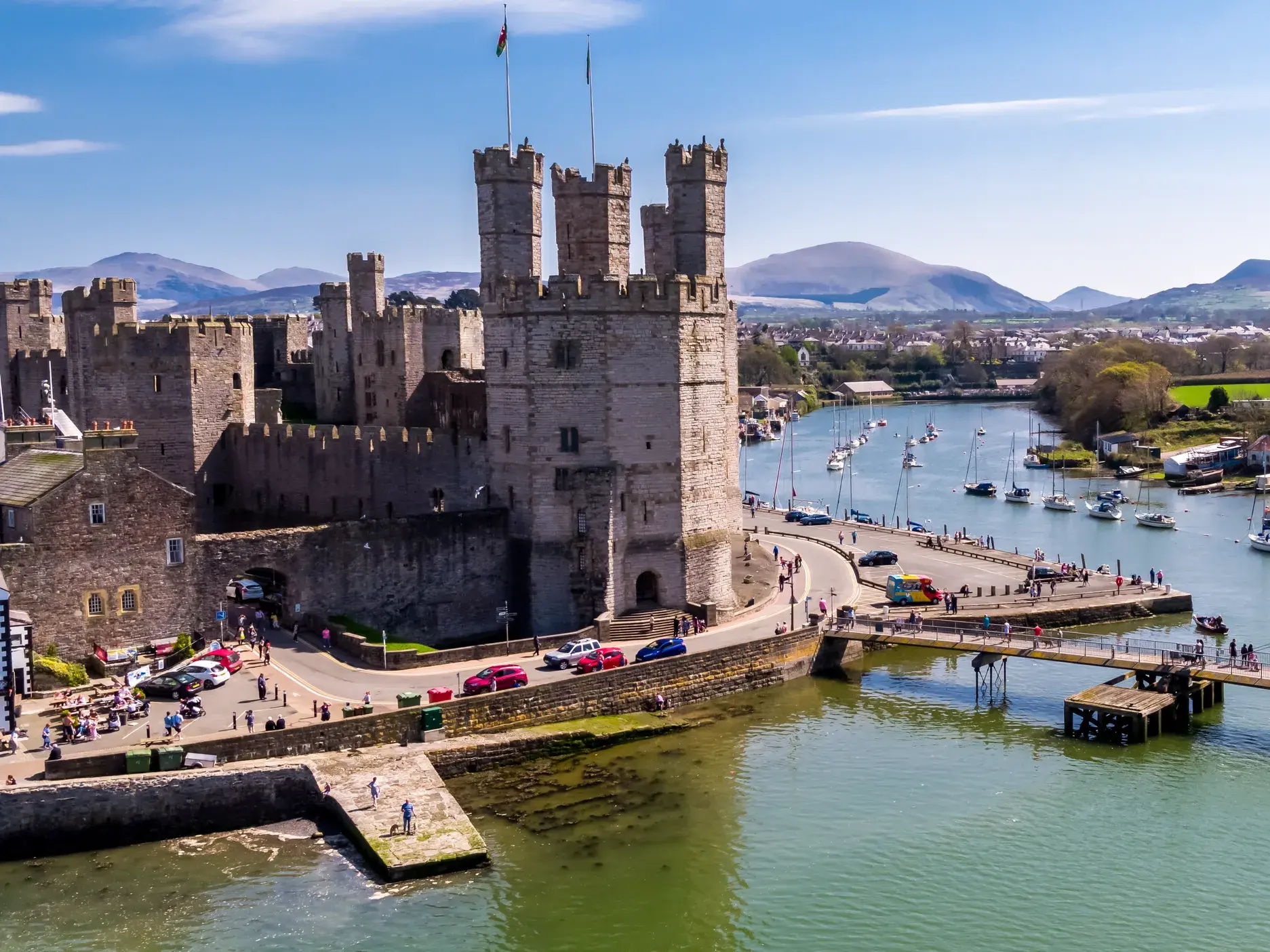 Luftaufnahme des UNESCO-Welterbes Caernarfon Castle in Wales, gelegen am Fluss Seiont mit Booten vor der Bergkulisse.