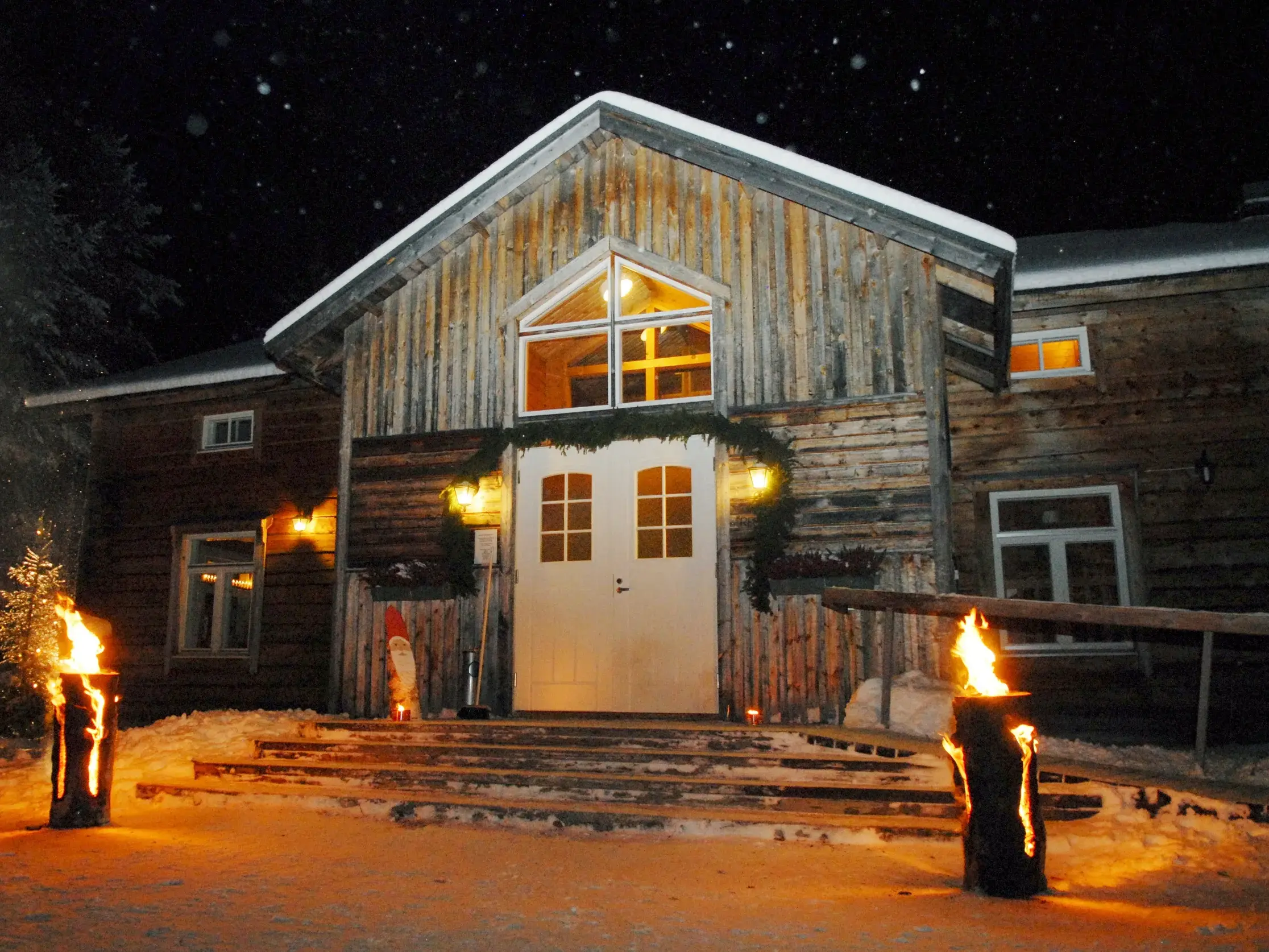 Nächtlicher Eingang der hölzernen Saija Lodge in Finnisch Lappland, beleuchtet von zwei lodernden Baumstamm-Fackeln im Schnee.