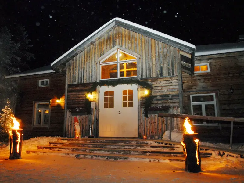 Nächtlicher Eingang der hölzernen Saija Lodge in Finnisch Lappland, beleuchtet von zwei lodernden Baumstamm-Fackeln im Schnee.