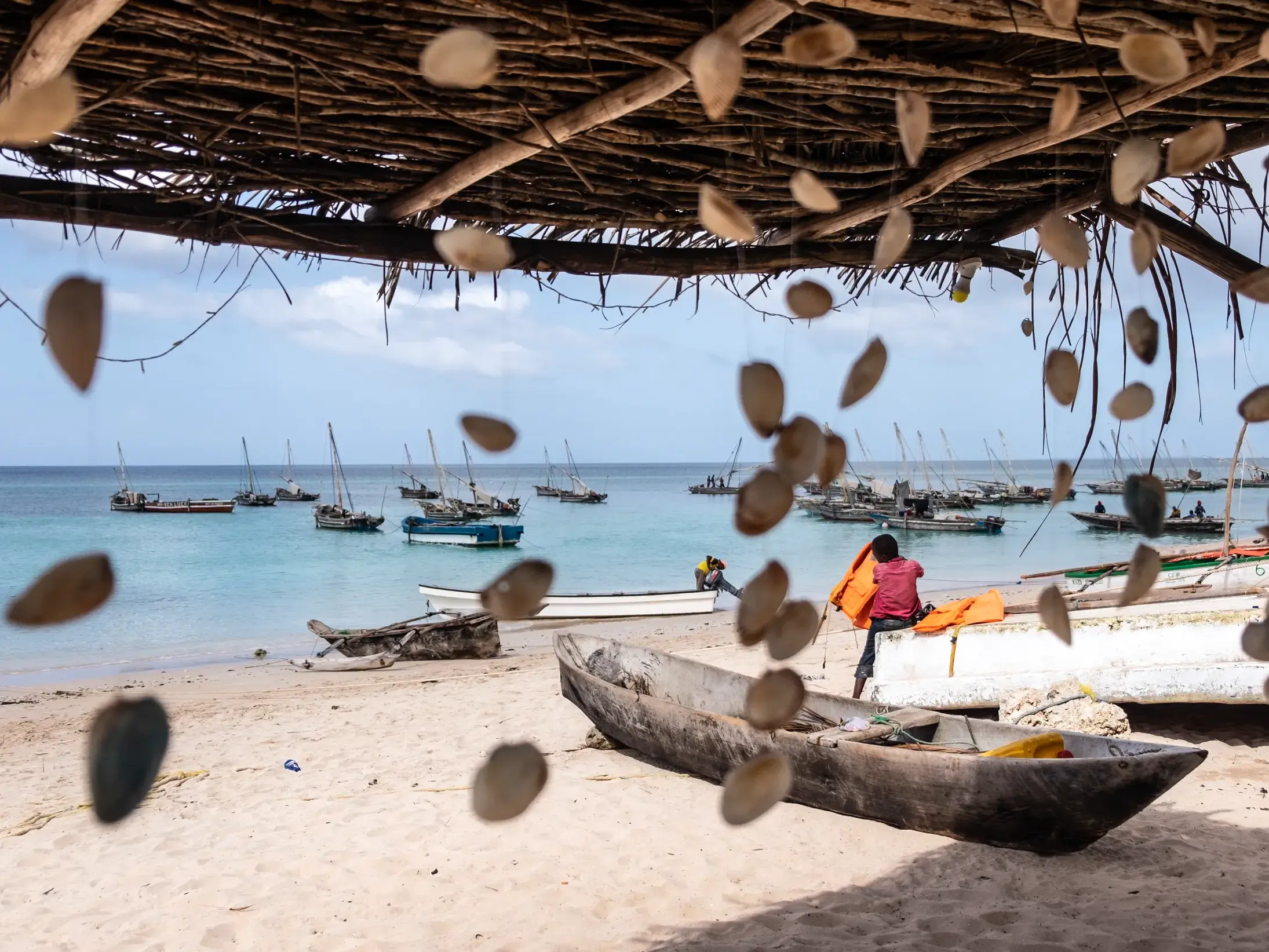 Blick durch einen Muschelvorhang auf den Sandstrand von Sansibar mit vielen traditionellen Dhaus im türkisen Meer.