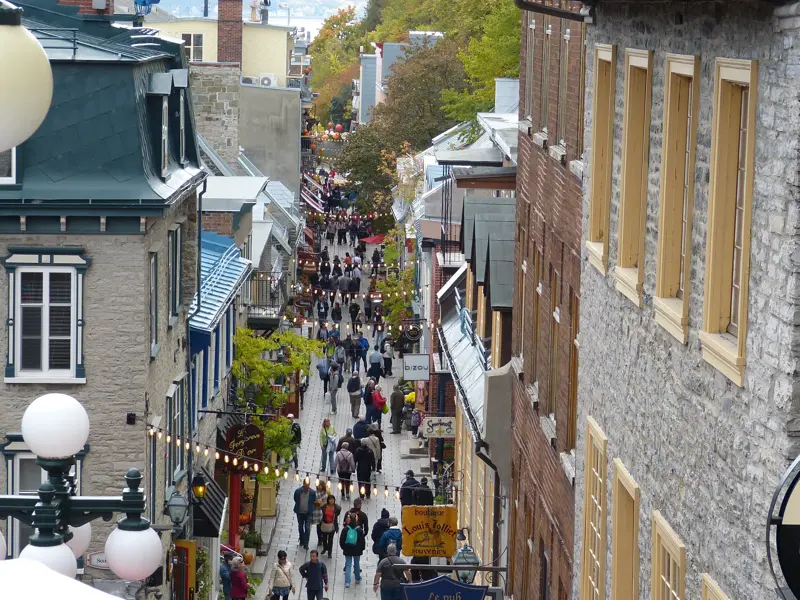 Menschen flanieren durch eine enge Gasse mit Steinhäusern und Lichterketten in der historischen Altstadt von Québec-Stadt.