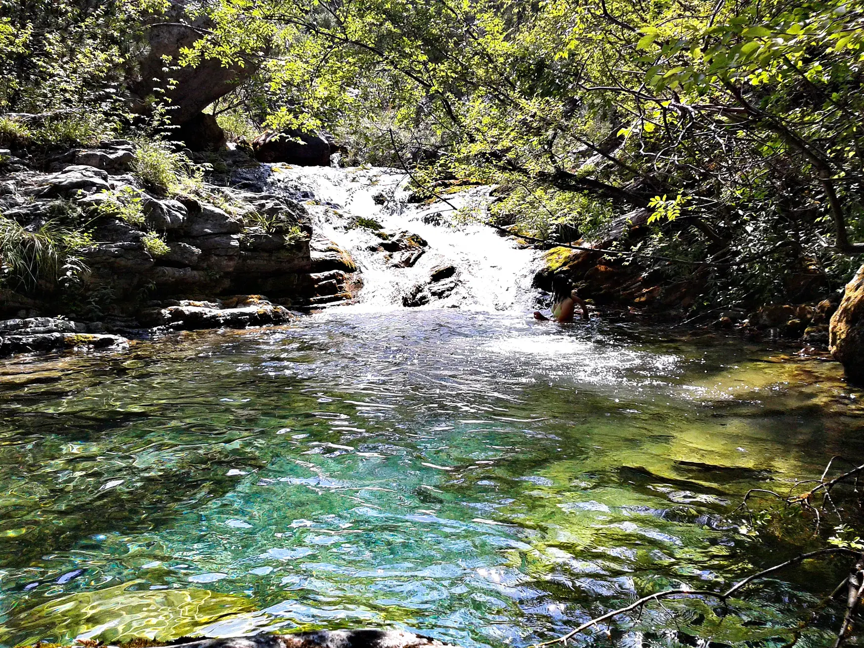Eine Person erfrischt sich in einem türkisfarbenen Gumpen eines Bergbachs in der sonnigen Enipeas-Schlucht in Griechenland.