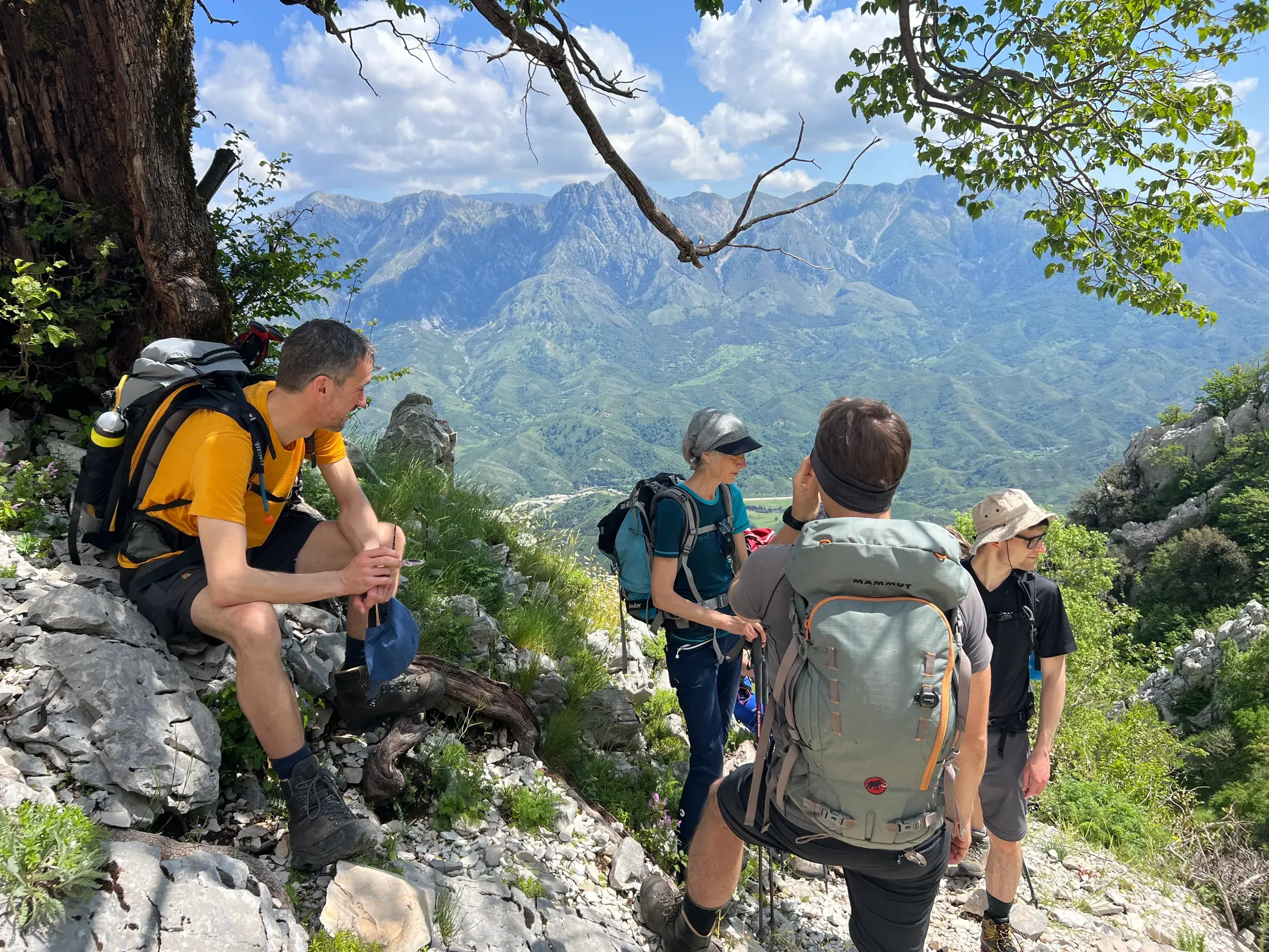 Eine kleine Wandergruppe mit Rucksäcken rastet auf einem steinigen Pfad im albanischen Küstengebirge mit weitem Ausblick.