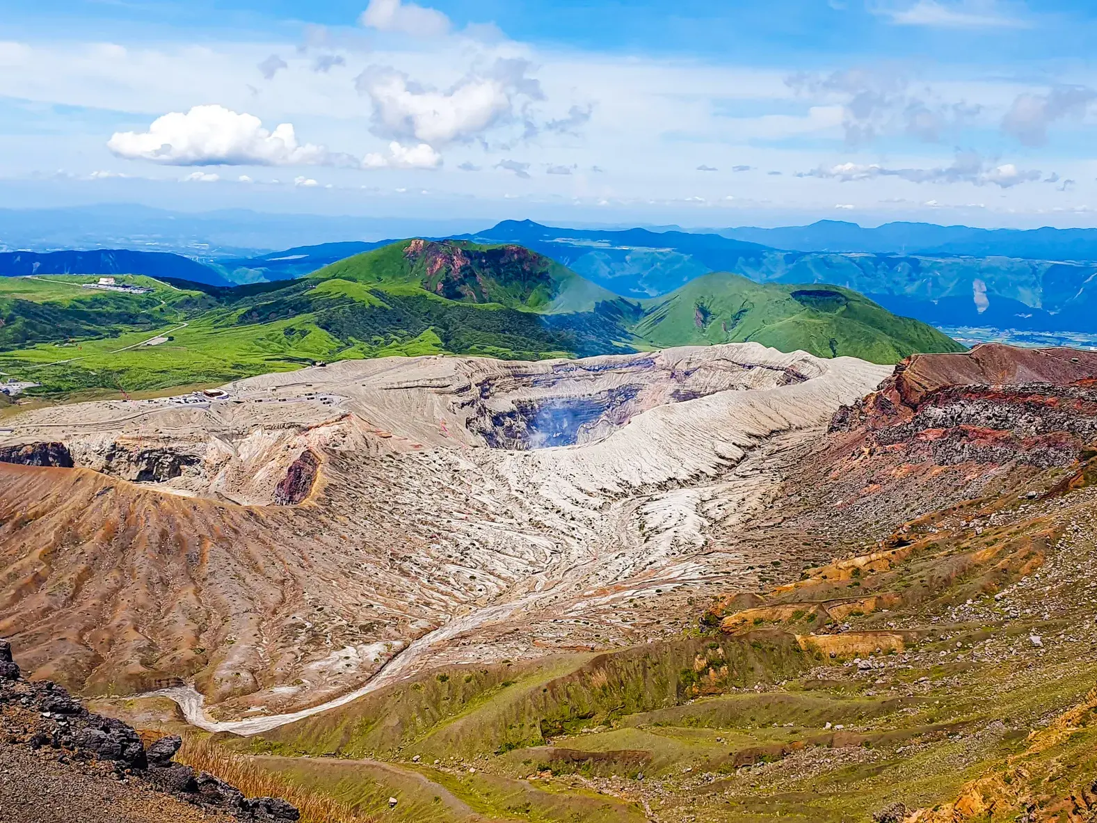 Weite Aussicht auf den aktiven Vulkankrater des Aso, umgeben von der grünen Berglandschaft des Aso-Kuju-Nationalparks, Japan.