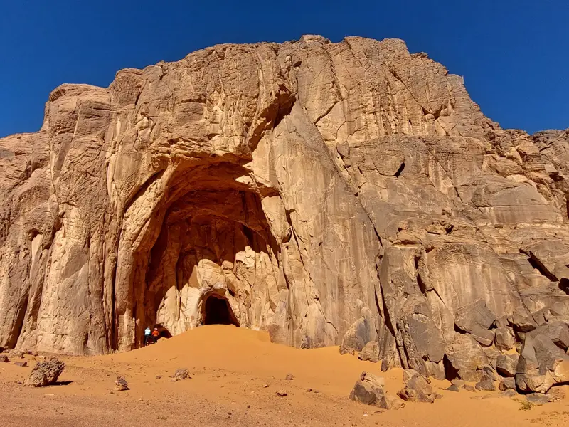 Zwei Wanderer vor einem monumentalen Höhleneingang in einer erodierten Sandsteinwand in der Wüste Algeriens.