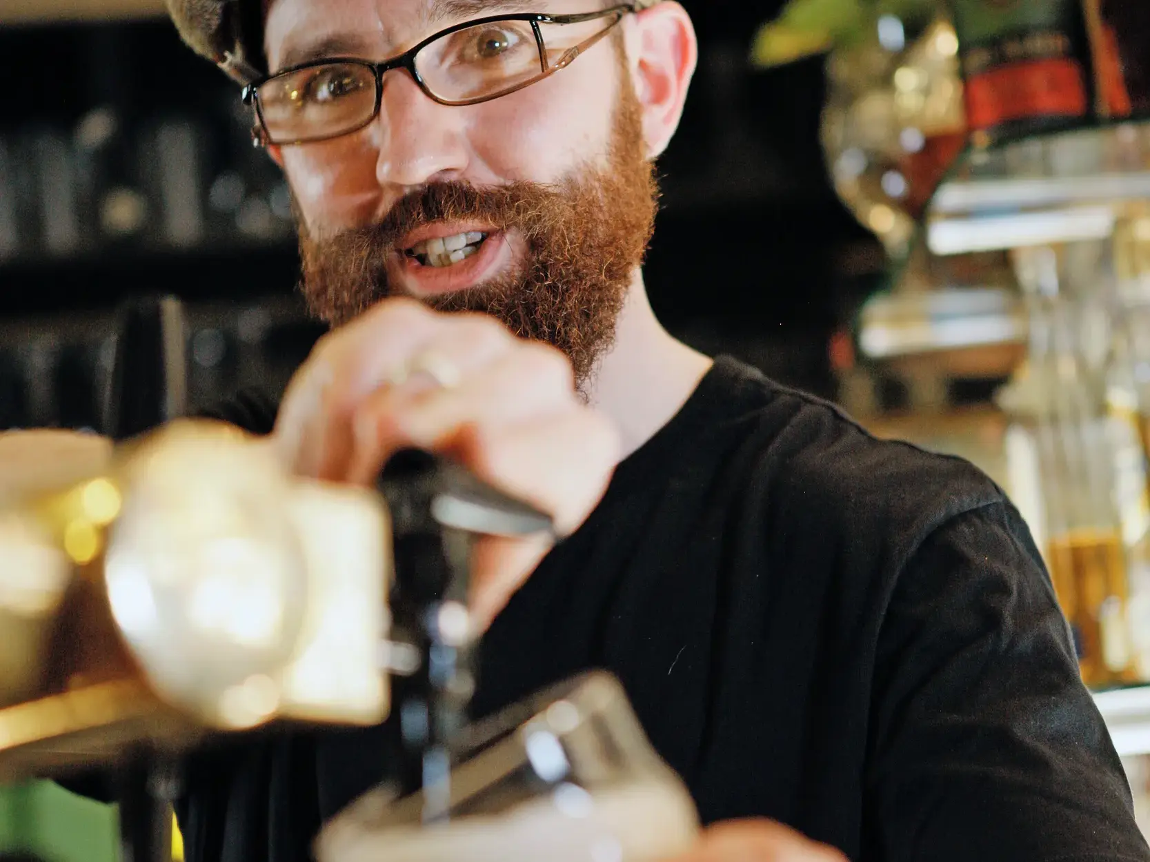 Ein bärtiger Barkeeper mit Brille zapft frisches Bier vom Fass in einem traditionellen Pub in Südengland.