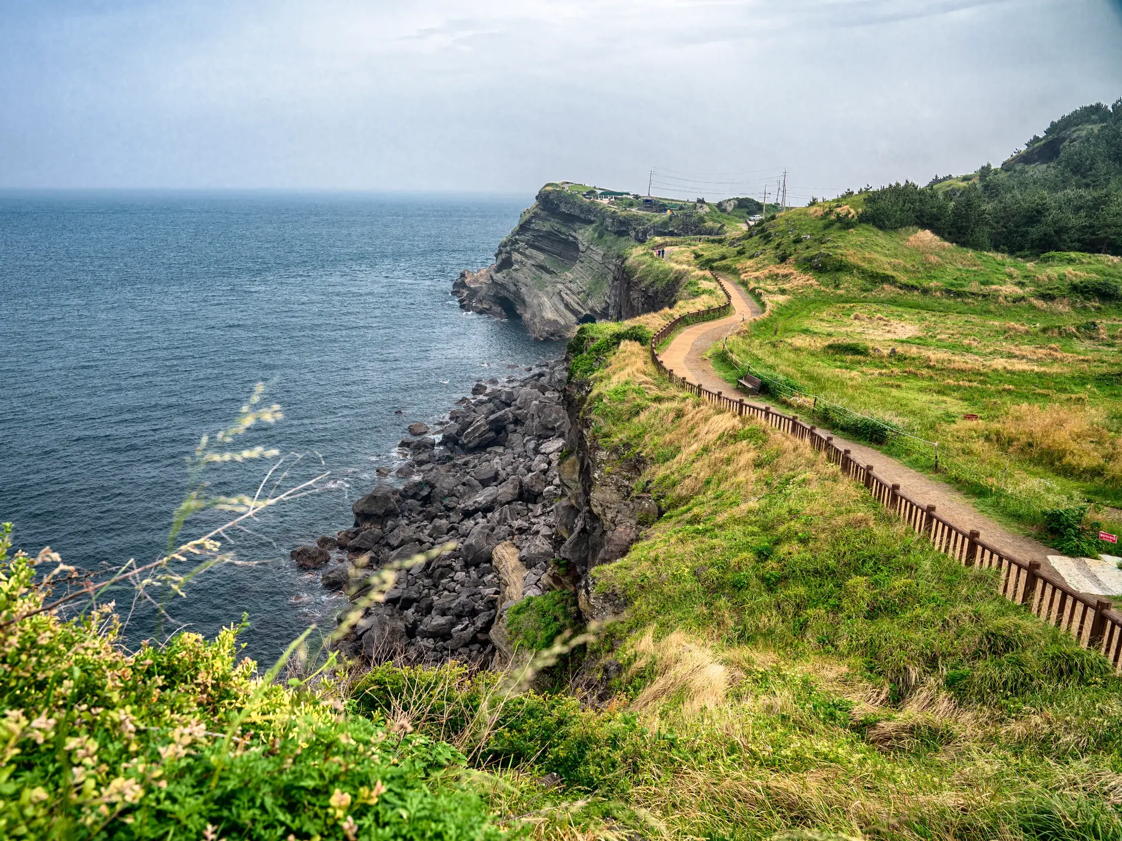 Ein malerischer Pfad führt entlang der grünen Vulkanküste der südkoreanischen Insel Jeju mit weitem Blick über das Meer.