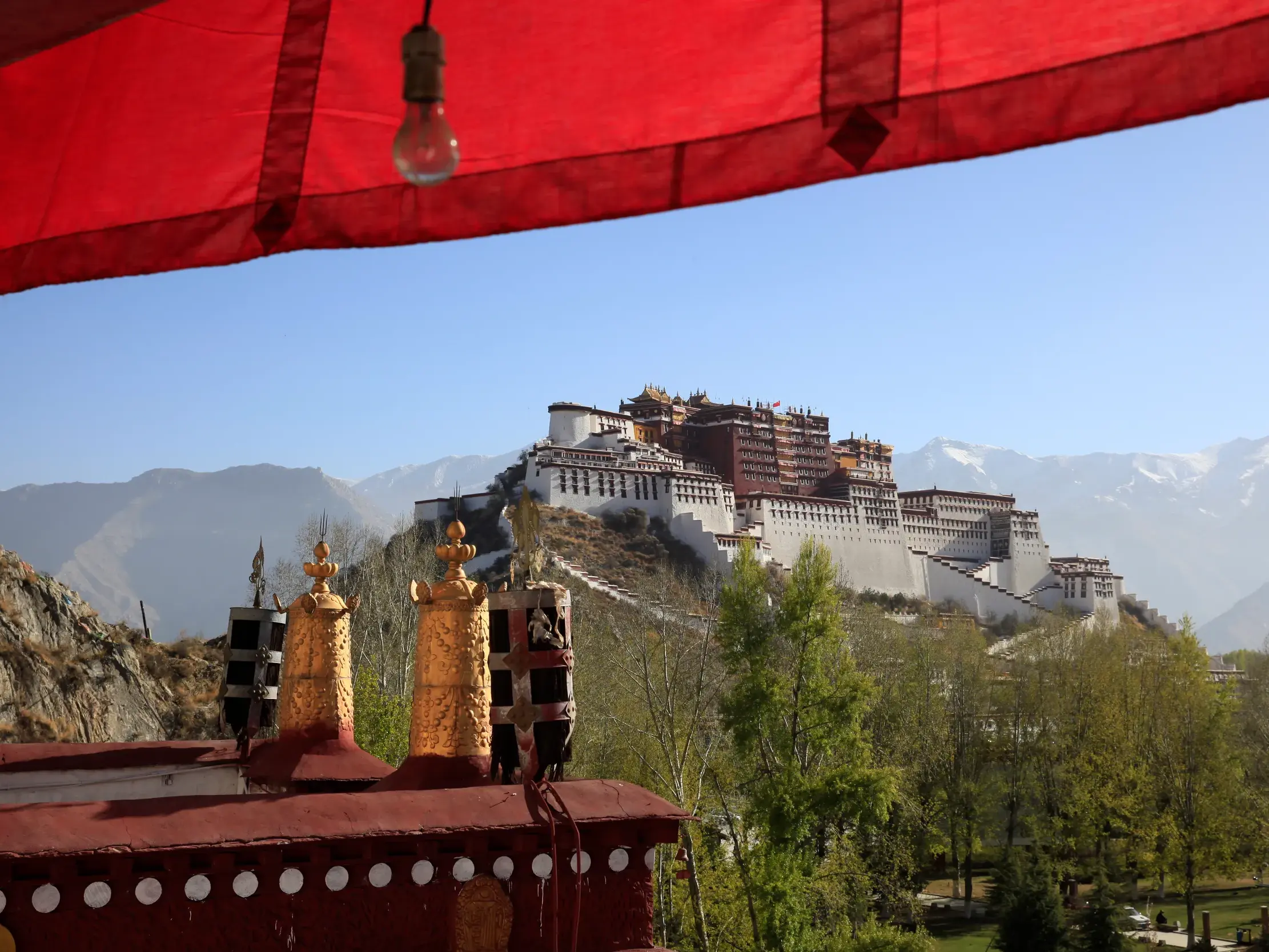 Der Potala thront erhaben auf einem Felsen in Lhasa, im Vordergrund ein Tempeldach, im Hintergrund die Berge Tibets.