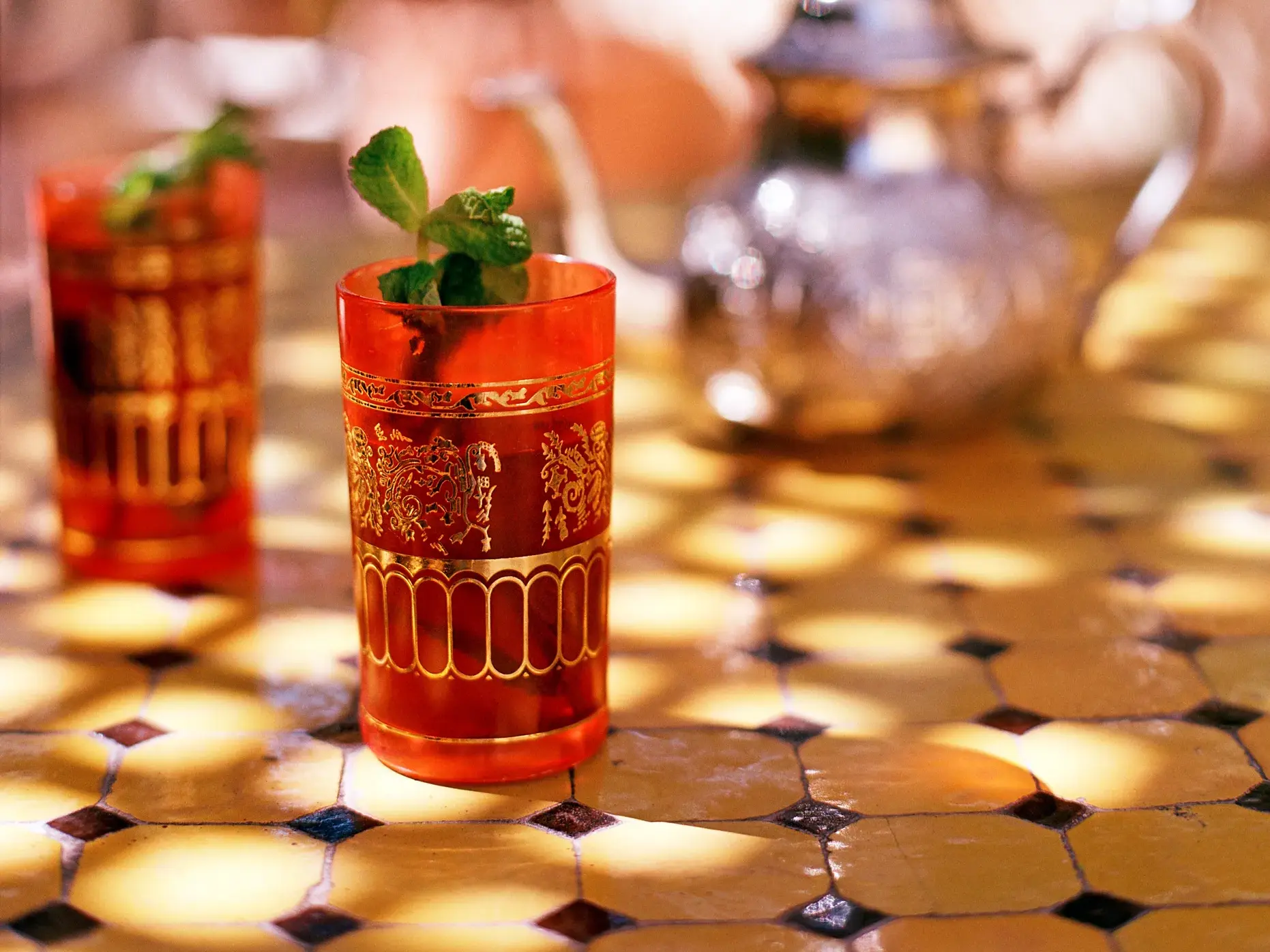 Zwei rote, goldverzierte Gläser mit marokkanischem Minztee auf einem Mosaiktisch im warmen Sonnenlicht in Marokko.