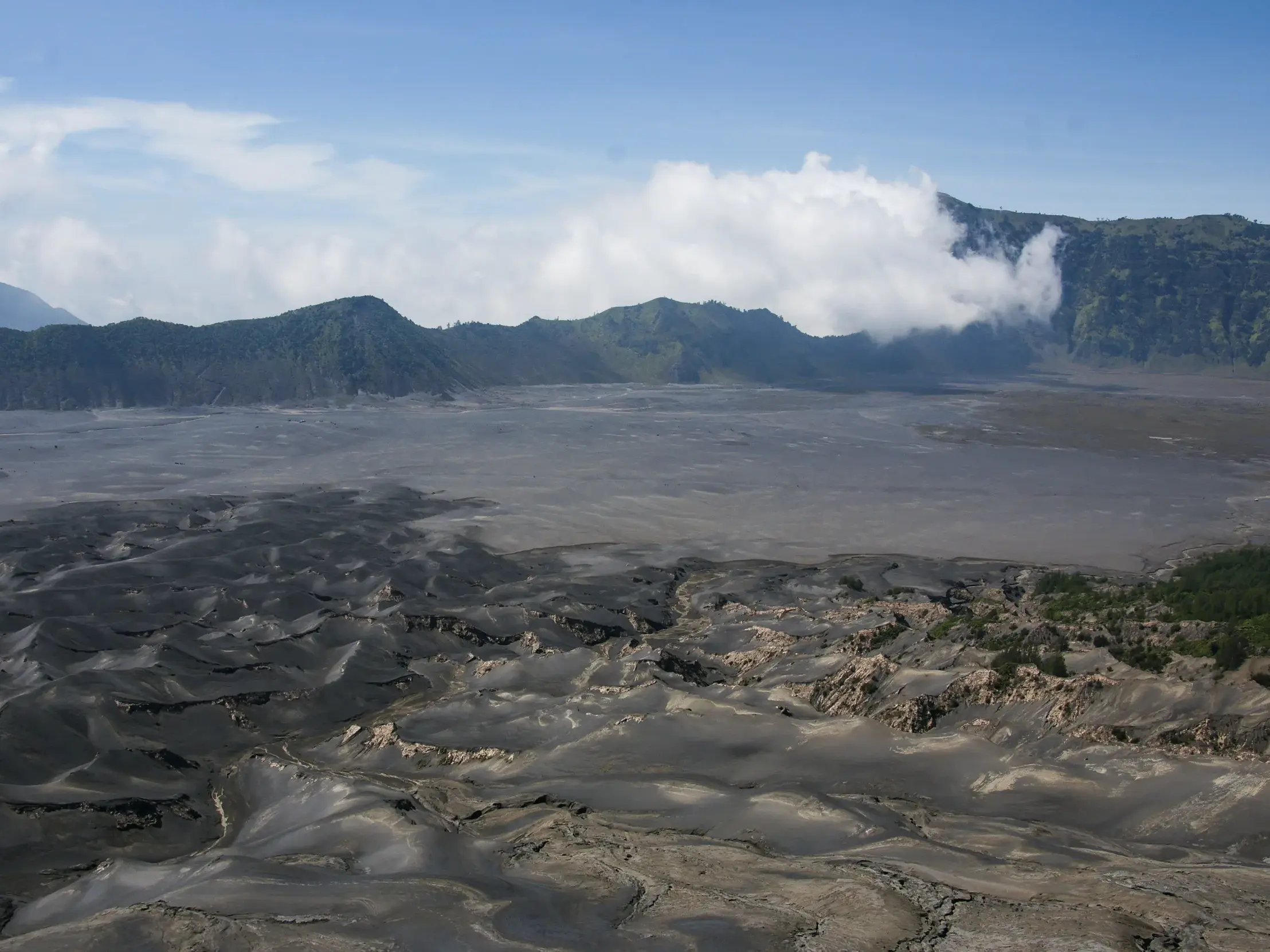 Karge Vulkanlandschaft des Bromo-Tengger-Semeru-Nationalparks auf Java mit dunklem Vulkansand und grünen Bergen im Hintergrund.