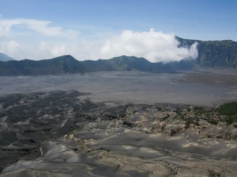 Karge Vulkanlandschaft des Bromo-Tengger-Semeru-Nationalparks auf Java mit dunklem Vulkansand und grünen Bergen im Hintergrund.