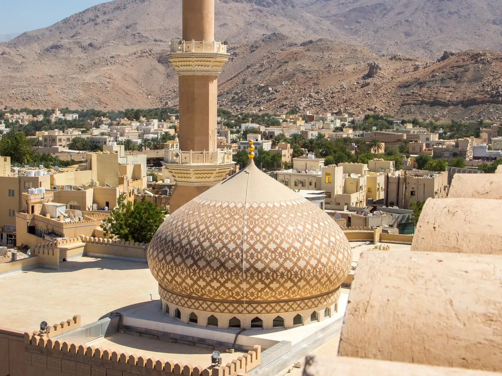 Blick von der Festung Nizwa auf einer Studiosusreise über die Dächer der Stadt, eine Moscheekuppel und das Hadschargebirge.