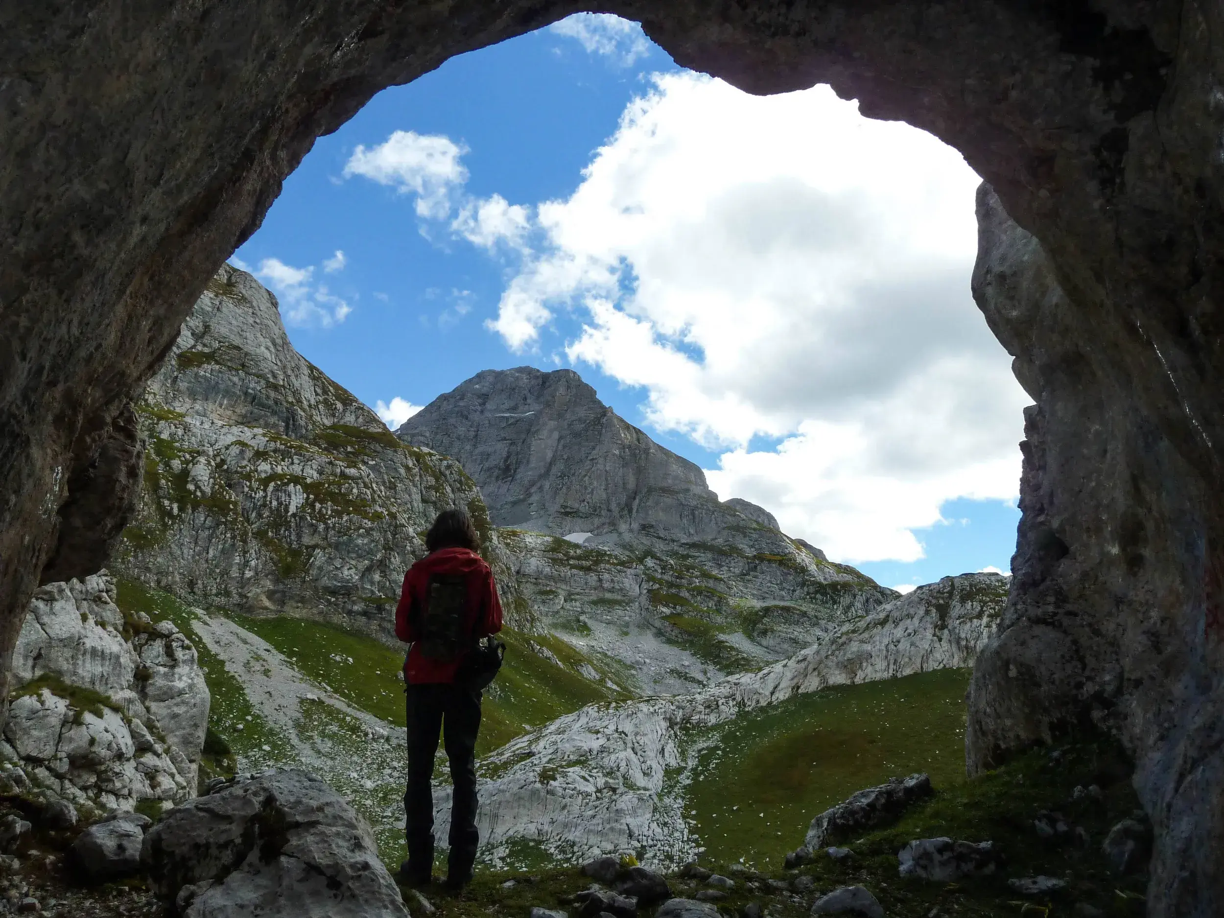 Eine Wanderin blickt aus einer Felshöhle auf die schroffen Gipfel des Prokletije-Gebirges auf dem Peaks of the Balkans Trail.