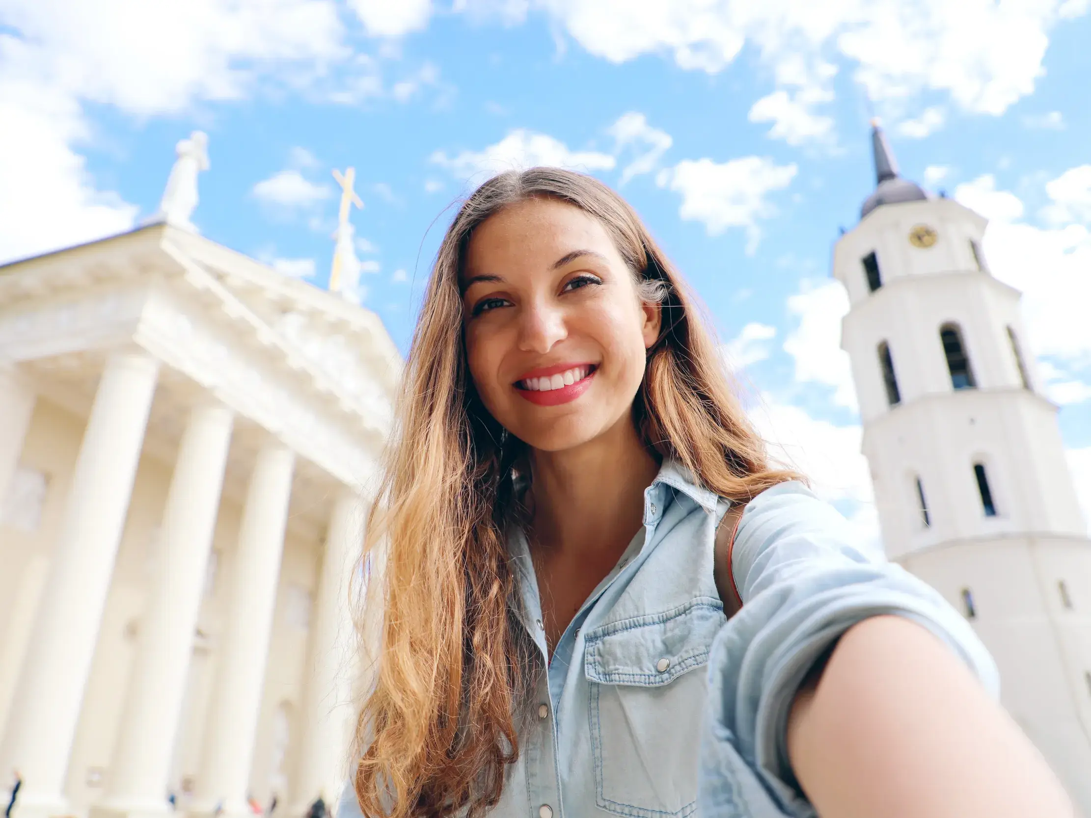 Eine junge Frau macht ein fröhliches Selfie vor der strahlend weißen Kathedrale von Vilnius und ihrem Glockenturm.