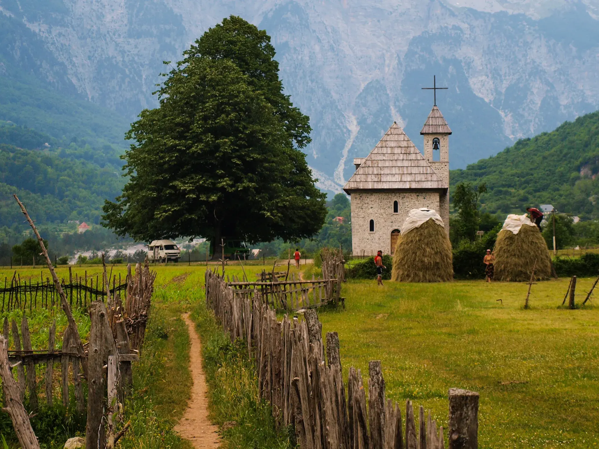 Die Kirche von Theth, Wahrzeichen des Bergdorfs, mit Heuhaufen auf einer Wiese vor der Bergkulisse der albanischen Alpen.
